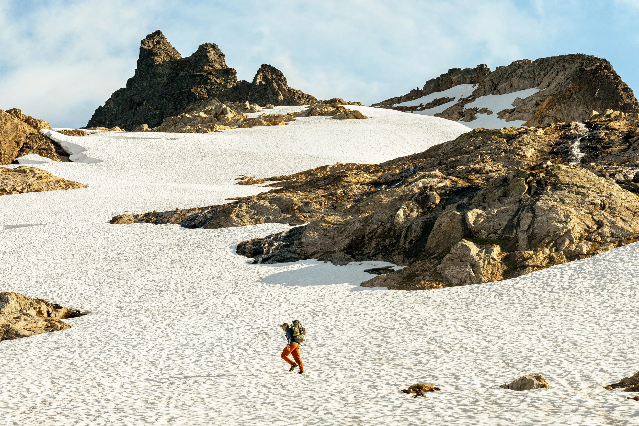 A person in orange pants and a blue jacket hiking across a snow-covered mountain landscape with rocky peaks in the background.