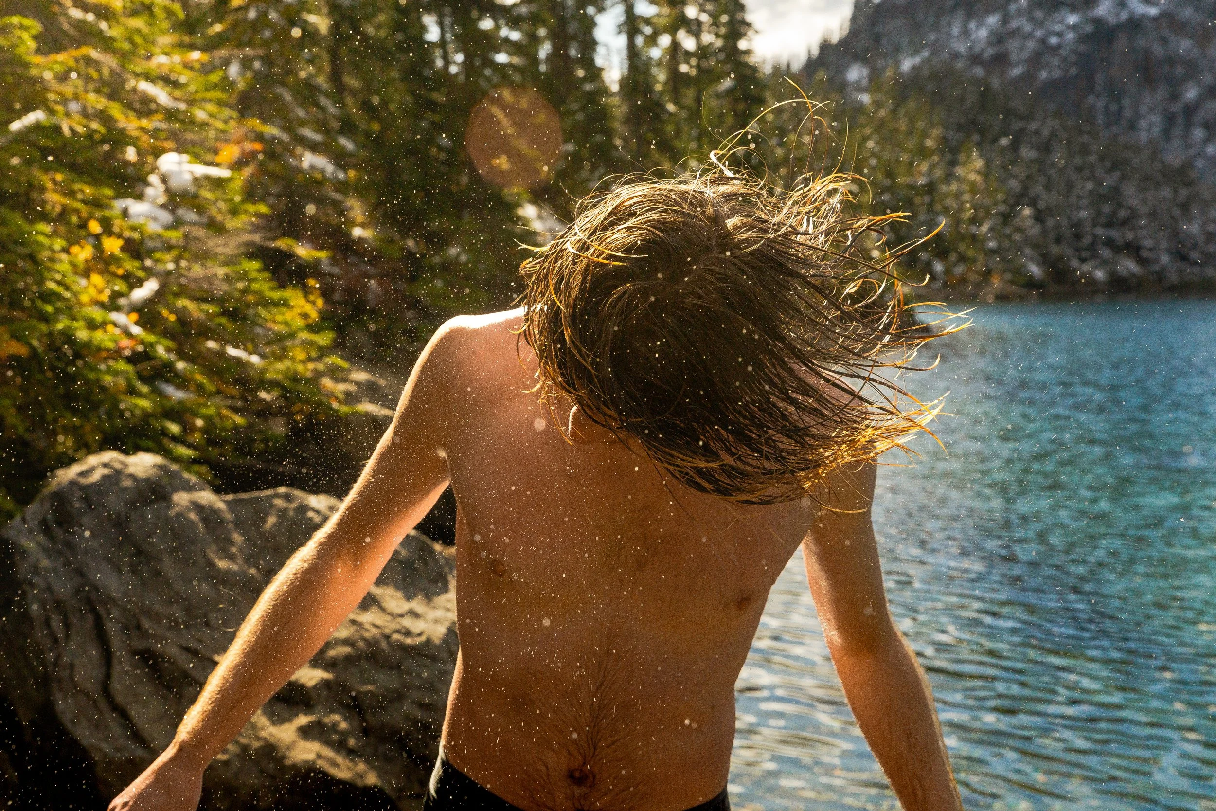 A shirtless person with long hair is flipping their hair in the water at a natural outdoor setting with trees, rocks, and a lake, during sunlight.