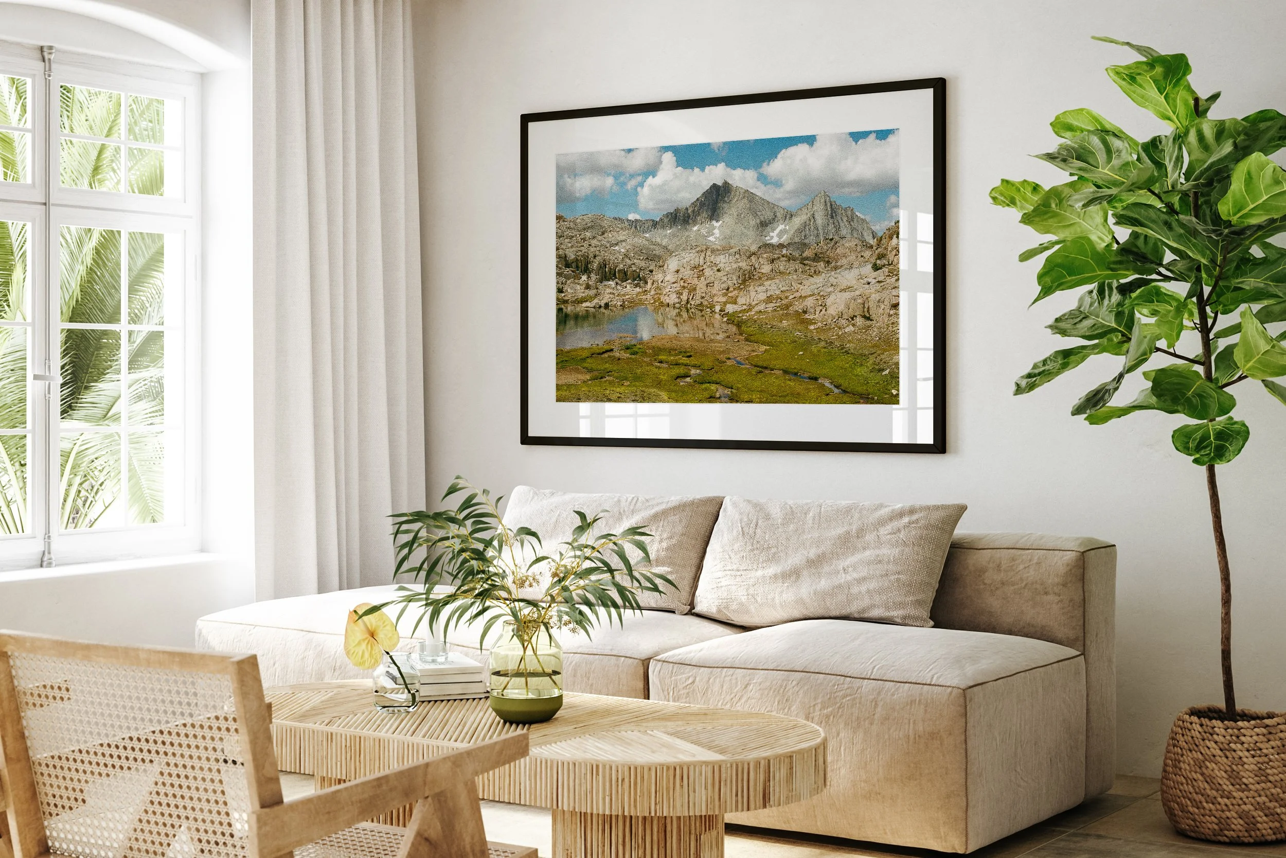 Living room with beige sofa, wooden coffee table with a green vase and plant, large window with white curtains, framed mountain landscape photograph on white wall, and a tall potted plant.