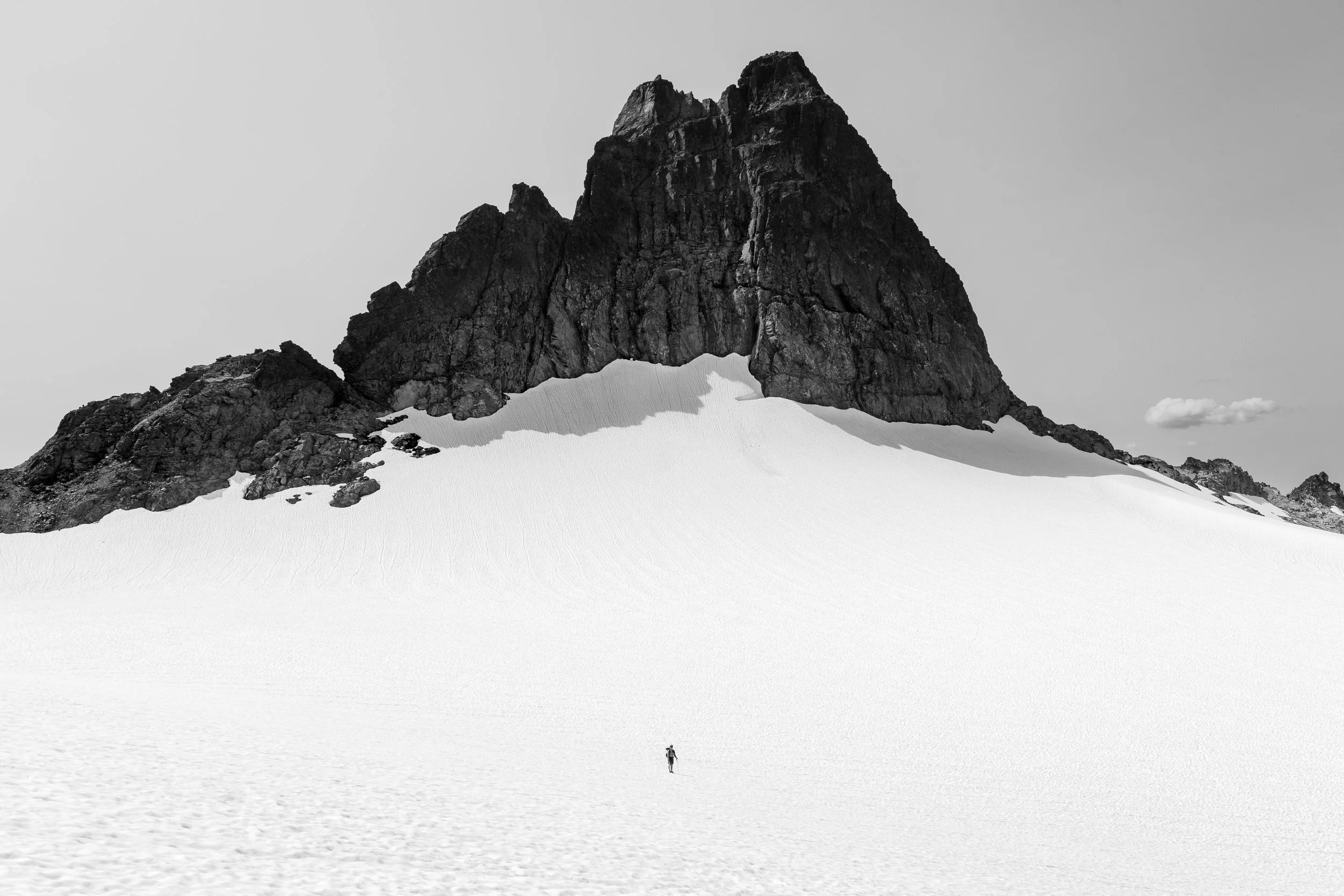 A lone hiker standing on snow-covered ground in front of a large dark rocky mountain with snow patches, under a partly cloudy sky.
