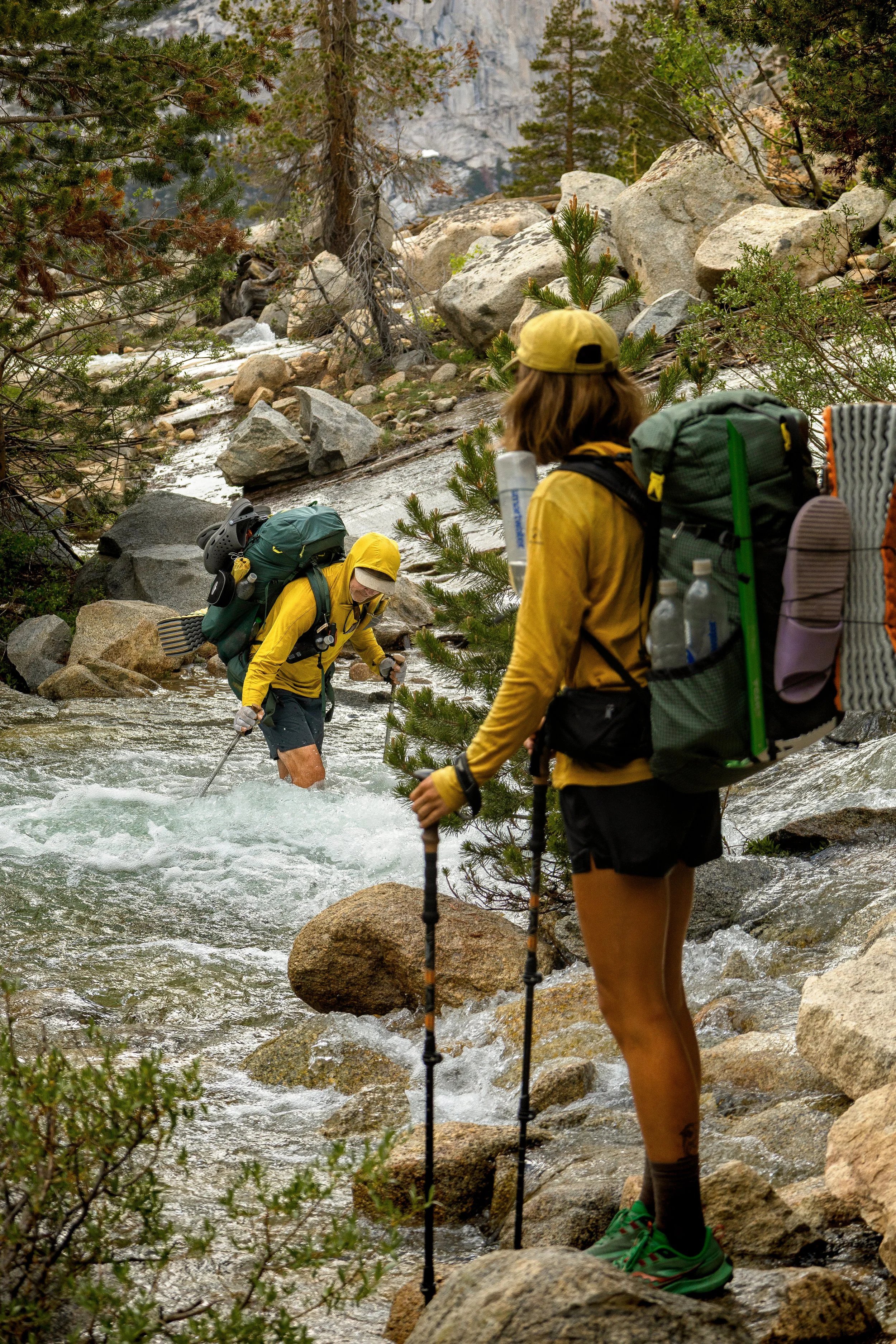 Two hikers crossing a rocky stream in a forested mountain area. One person is wearing a yellow hat and yellow jacket, holding trekking poles, and a large backpack. The second person, in a similar hat and jacket, is partially visible, standing on a rock.