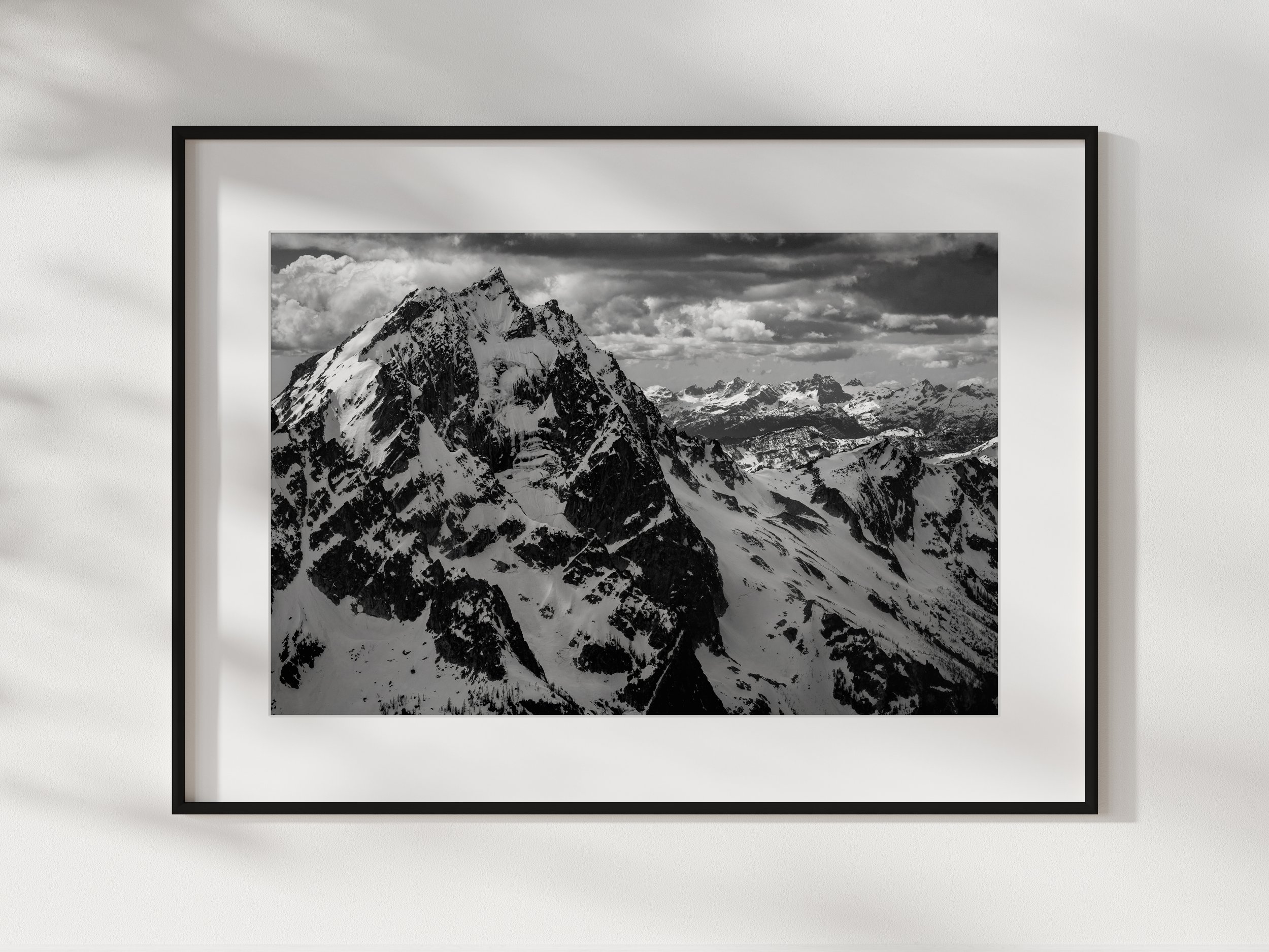 Black and white photograph of snow-covered mountain peaks under a cloudy sky, framed on a white wall.