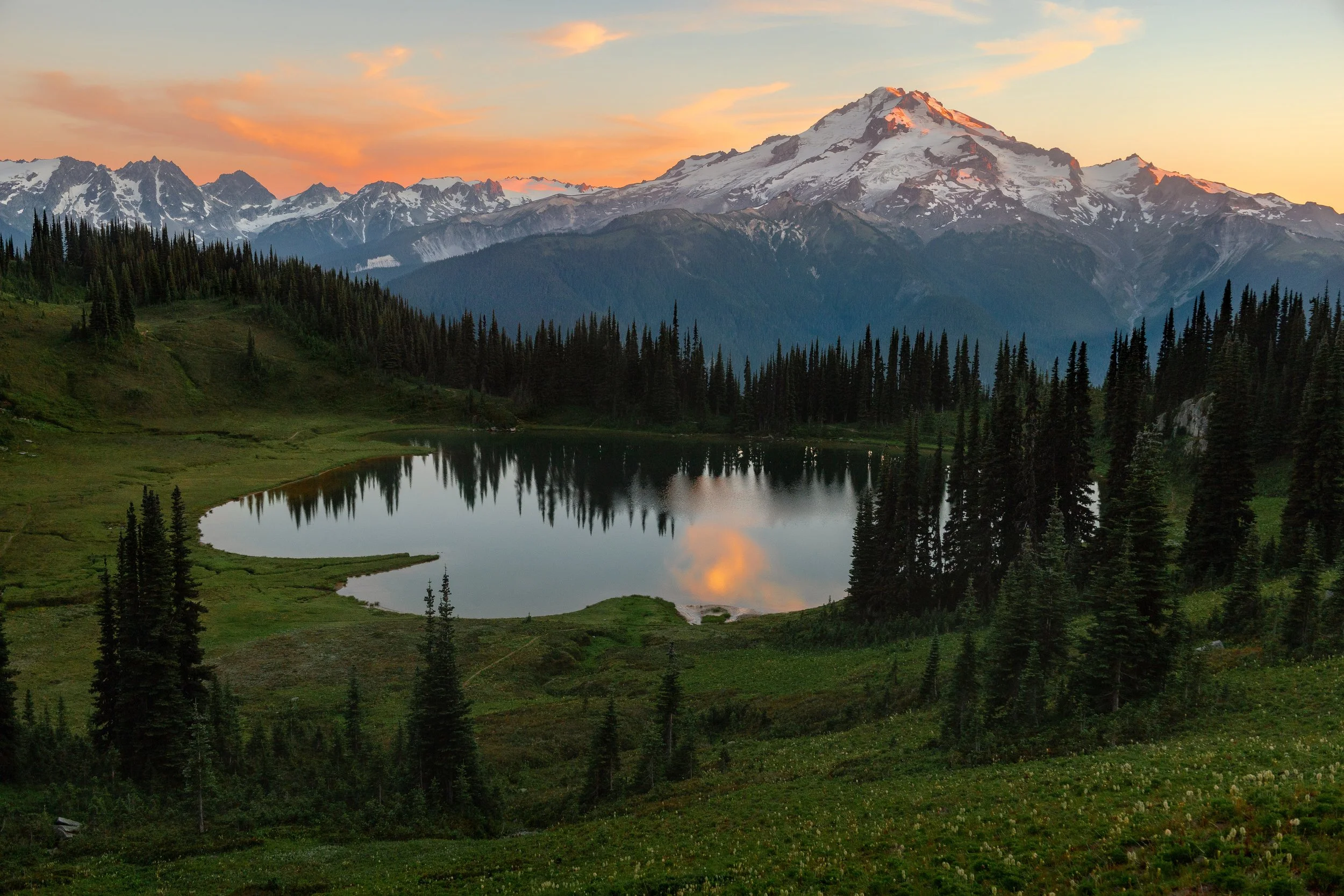 A serene mountain landscape featuring a snow-capped peak in the background, a reflective lake in the center, and dense pine trees surrounding the lake at sunset with orange and pink hues in the sky.