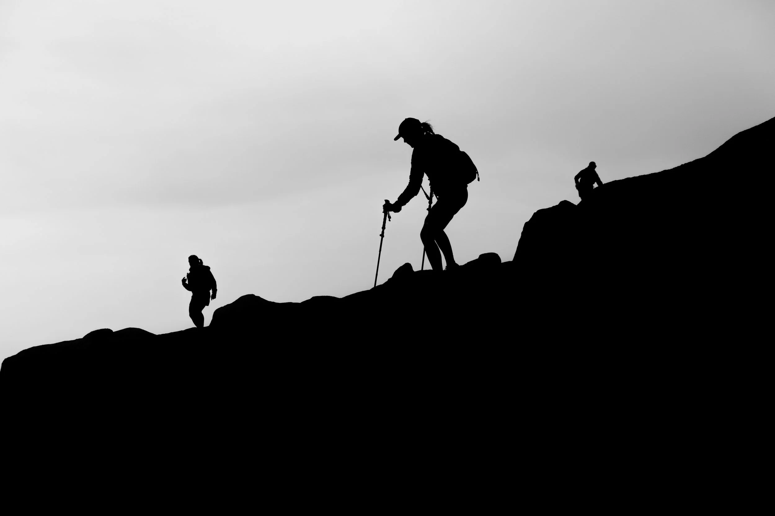 Silhouettes of people hiking up a mountain slope against a cloudy sky.