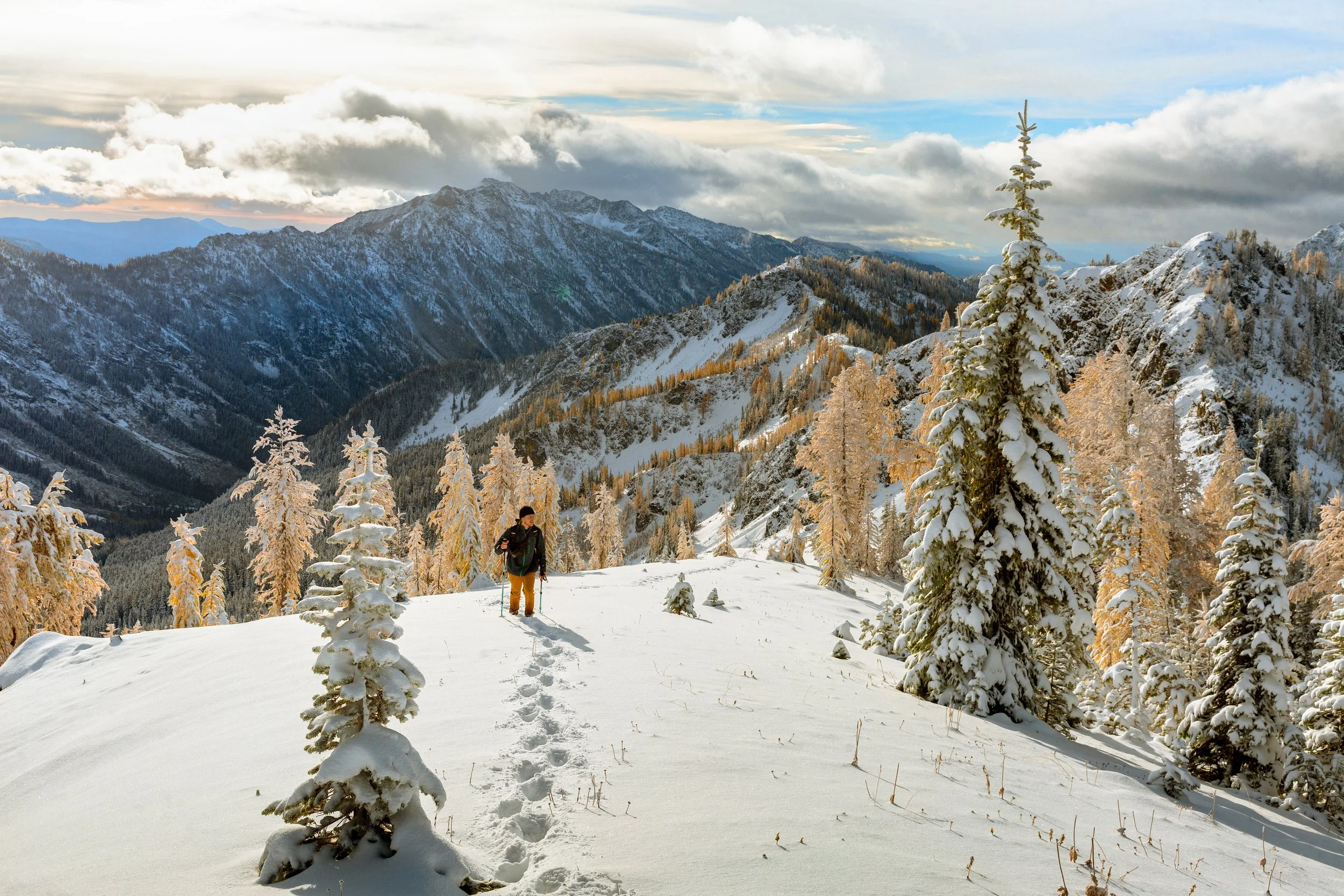 A person hiking in a snowy mountain landscape with snow-covered trees in the foreground, mountain ridges in the background, and a partly cloudy sky.