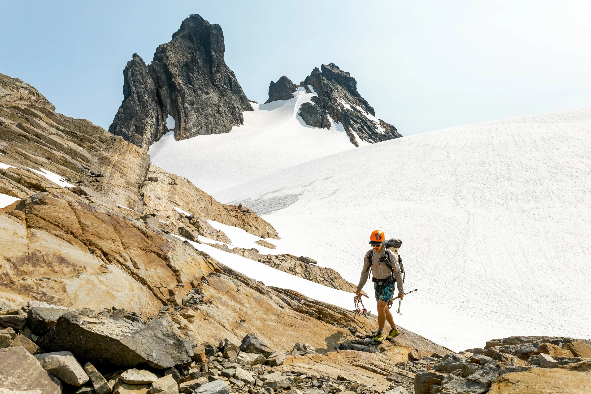 A mountain climber wearing an orange helmet and sunglasses ascending a rocky slope near snow-covered terrain with a steep mountain peak in the background.