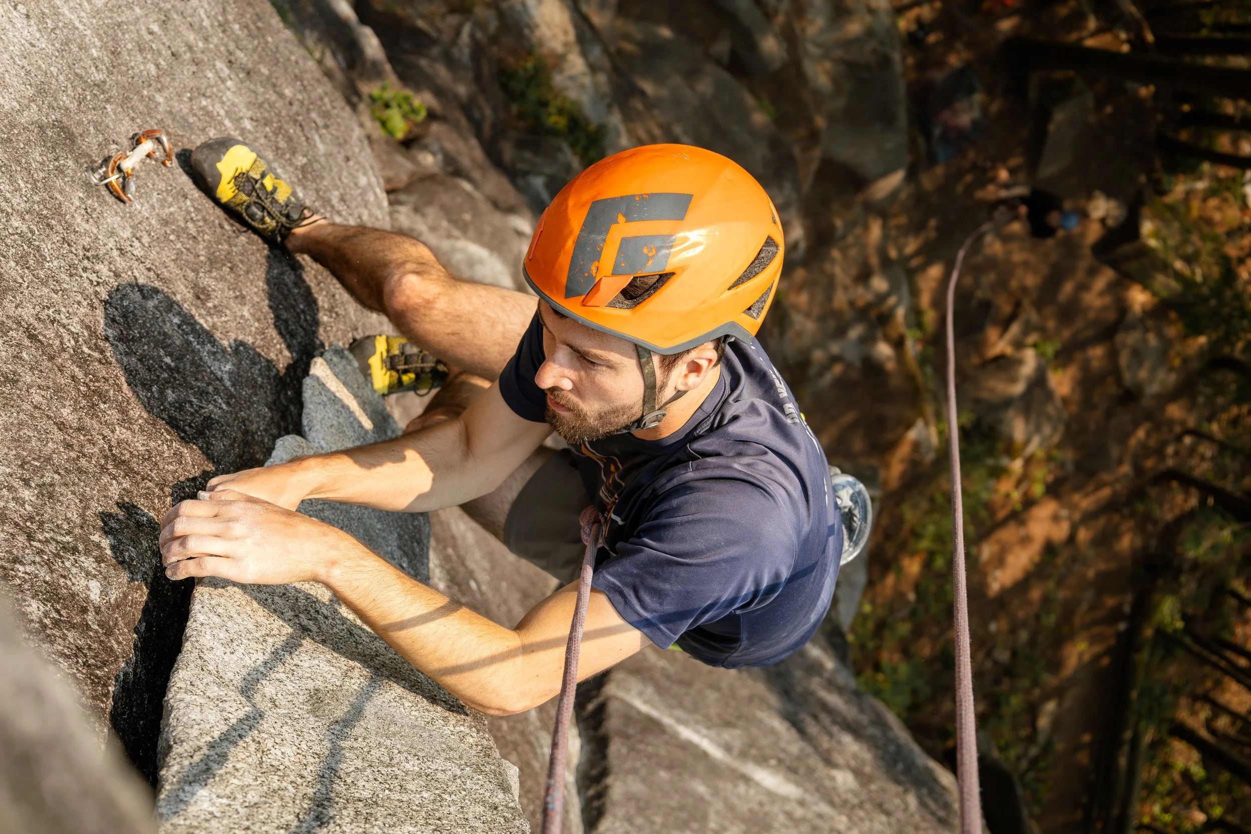 Man rock climbing on a steep rock face, wearing an orange helmet, black shirt, and climbing shoes, with a safety harness and rope, surrounded by trees and rocks.