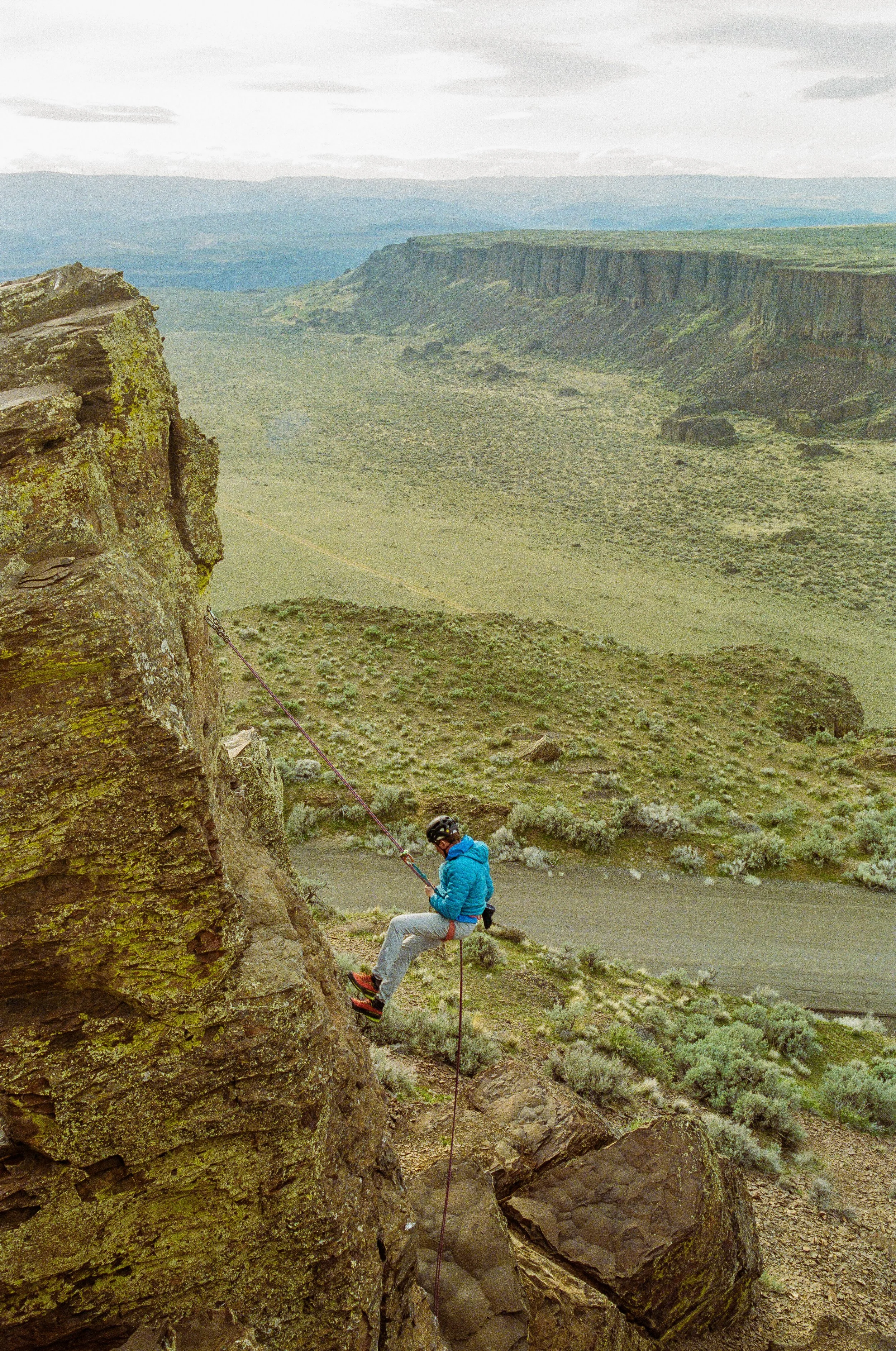 A person wearing a blue jacket, gray pants, red shoes, and a black helmet is sitting on a rocky ledge holding a climbing rope. The landscape features large rock formations, bushes, and distant cliffs under a cloudy sky.