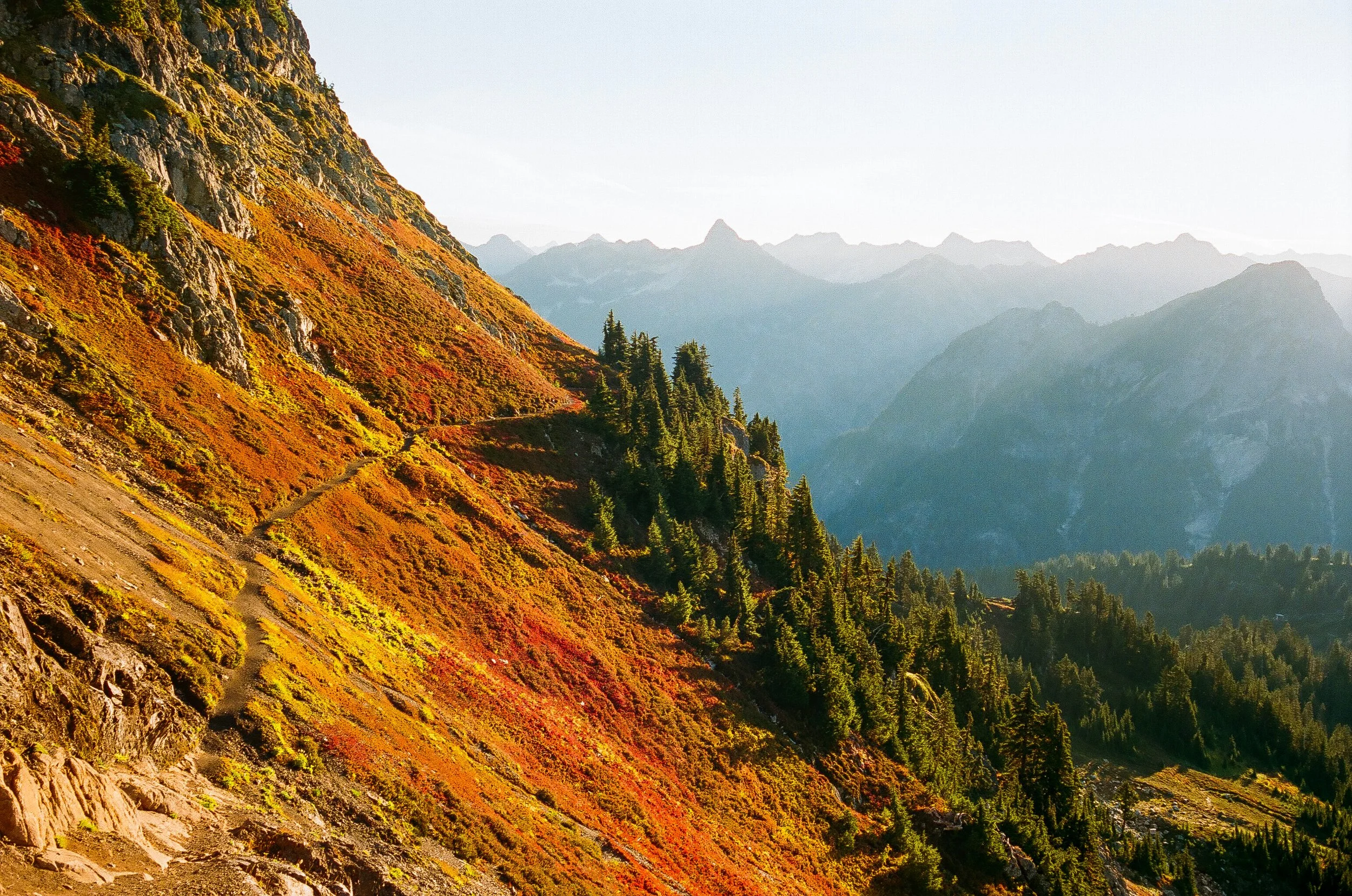 A mountain landscape with a steep, grassy slope covered in patches of orange, yellow, and green vegetation. In the background, rugged mountain peaks are visible under a clear sky, with the mountains fading into the distance.