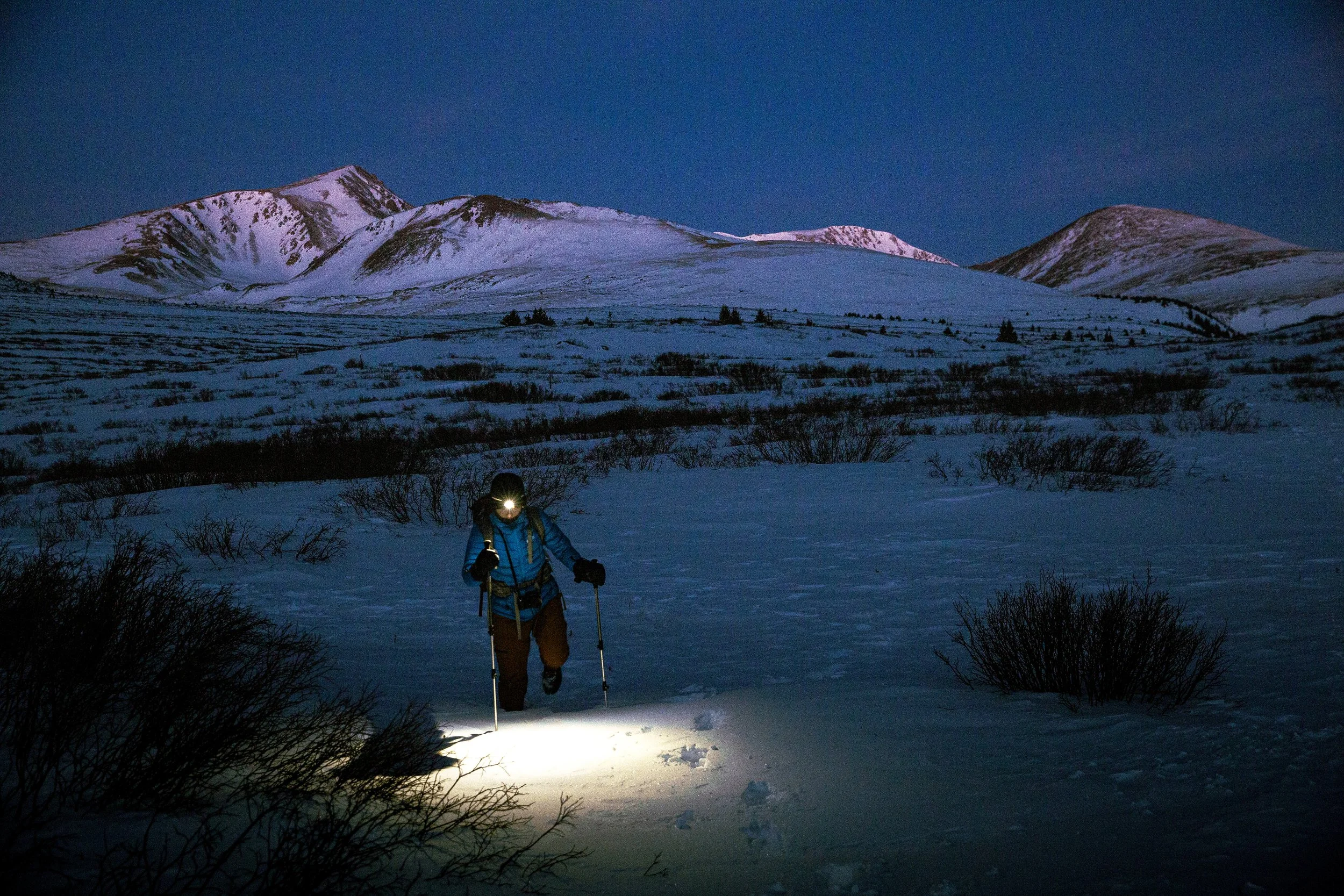A person dressed in outdoor winter gear, including a headlamp, hiking poles, and a jacket, is walking through snow at night in a mountainous area with snow-covered peaks in the background under a dark sky.