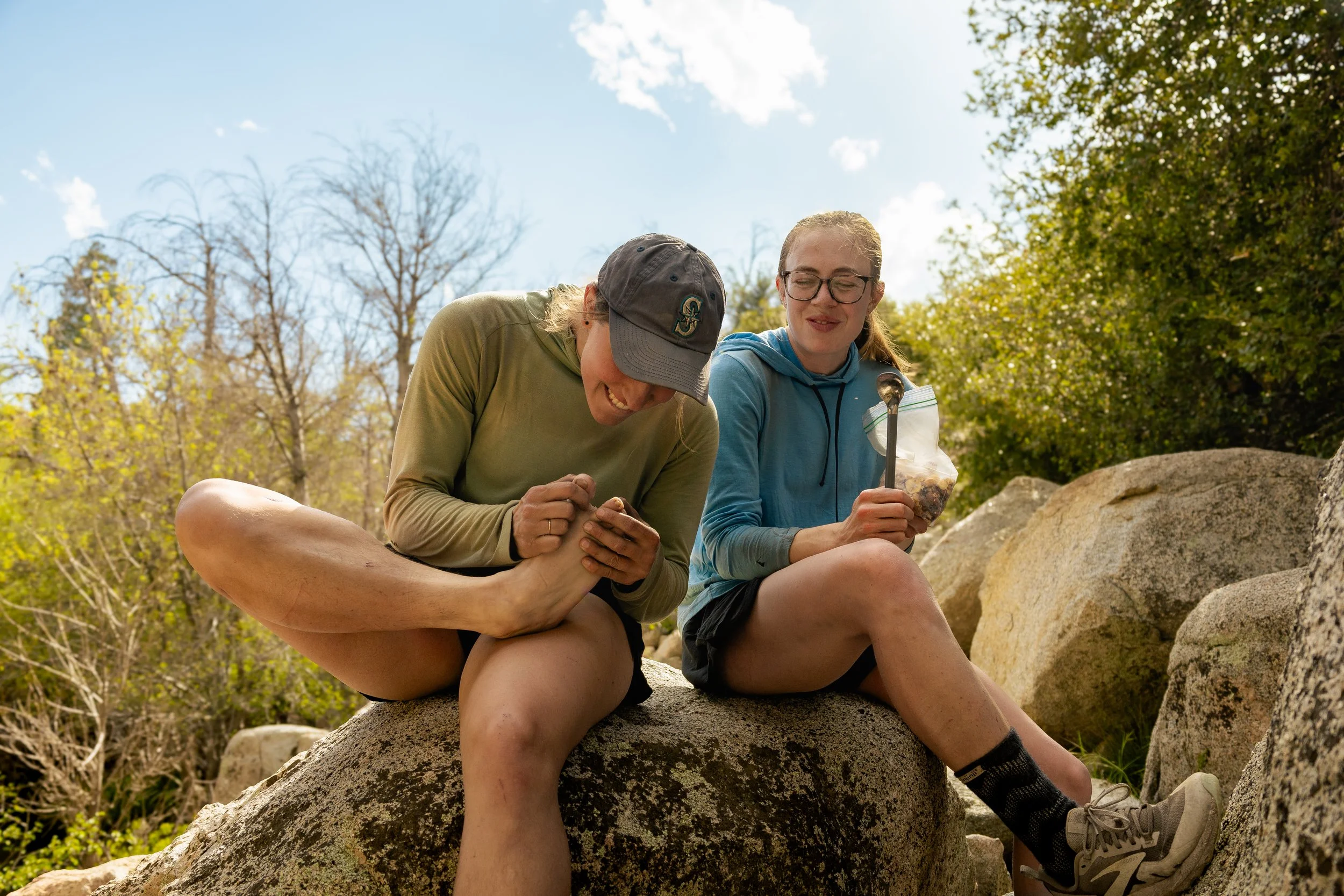 Two people sitting on large rocks outdoors, one laughing and holding a foot, the other holding a bag of trail mix, with trees and blue sky in the background.