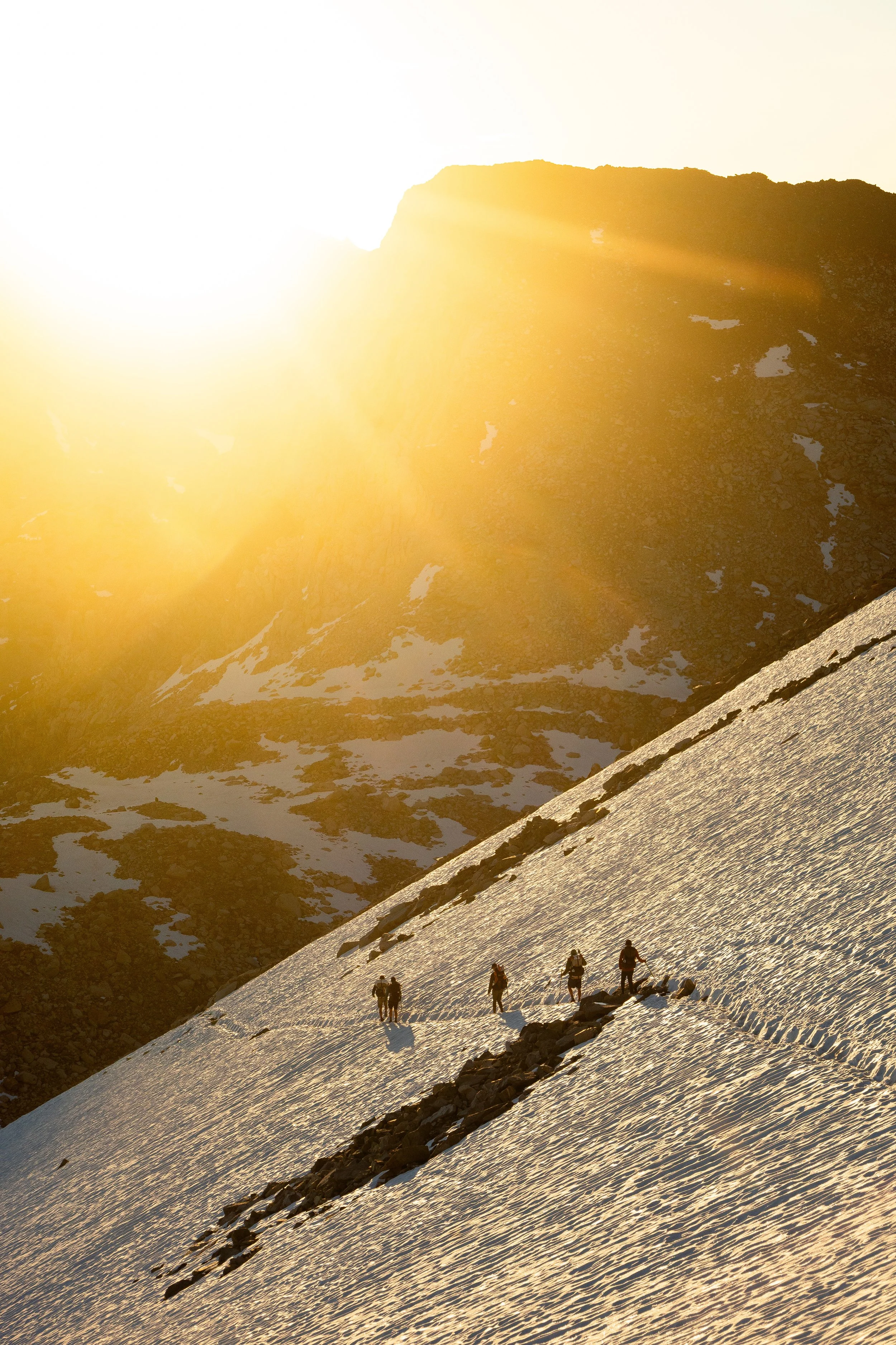 A group of hikers trekking across a snowy mountain slope during sunset, with mountain peaks in the background.