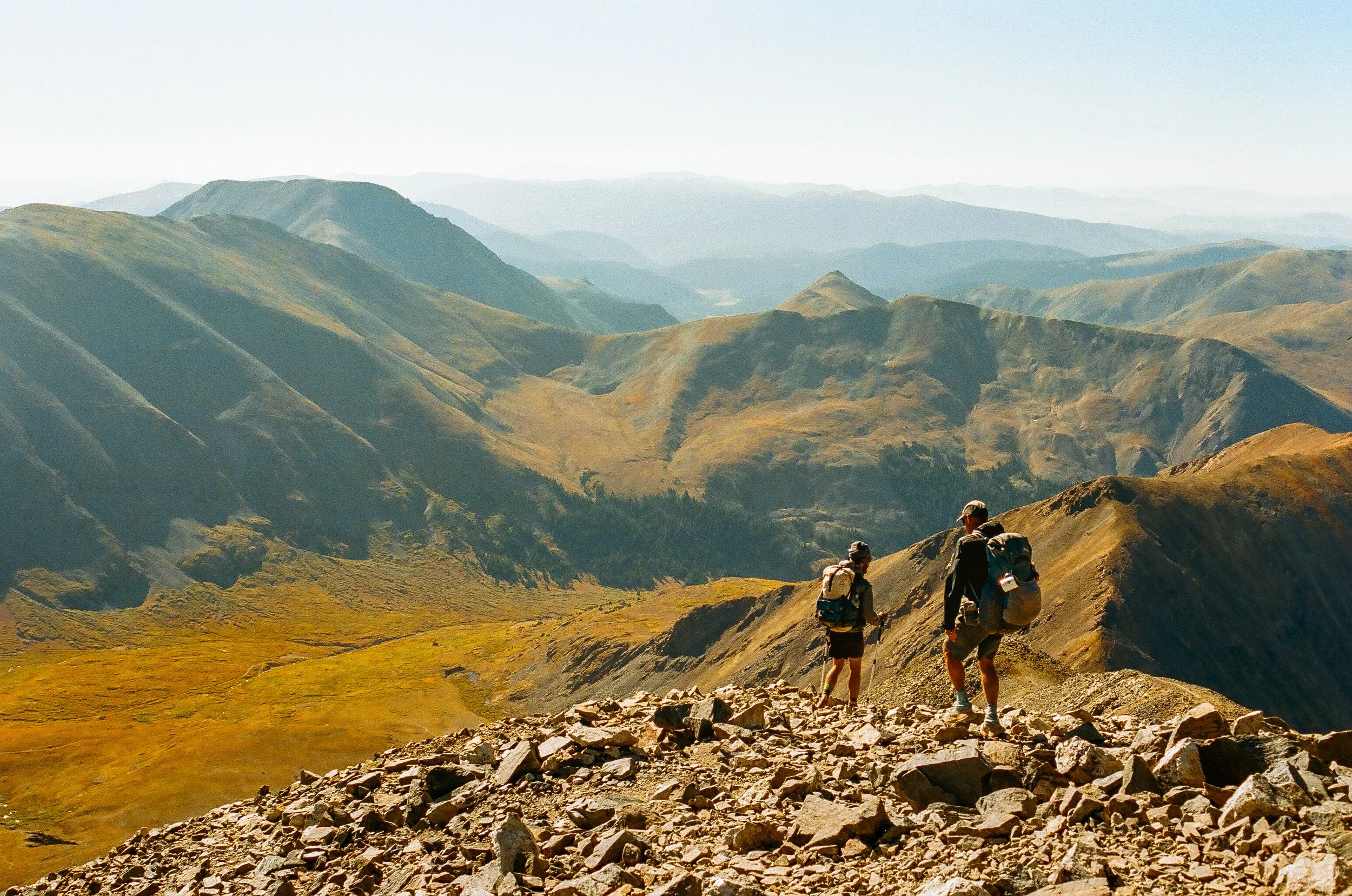 A group of hikers walking along a rugged mountain trail with rolling mountain ranges in the background under a clear sky.