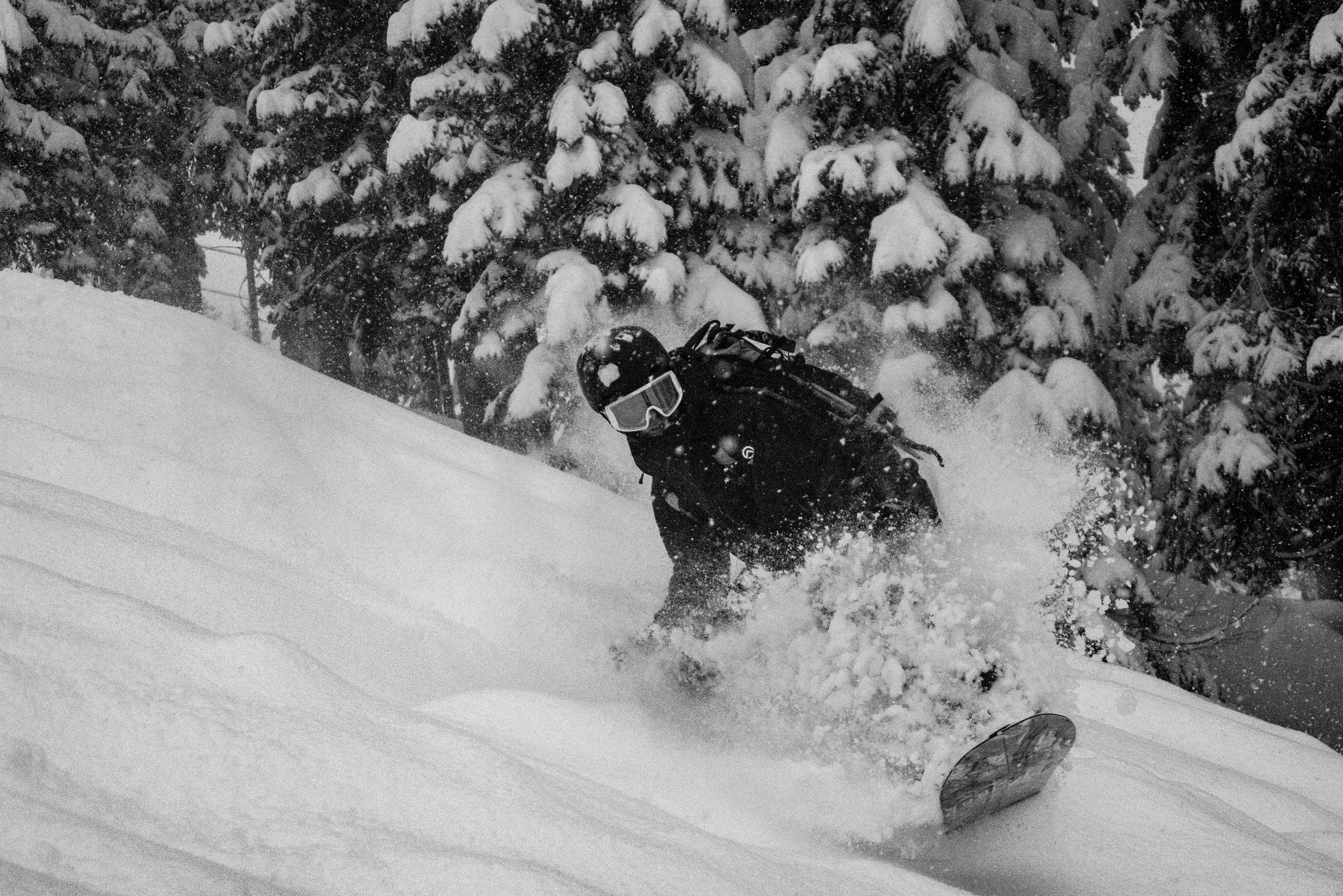 A skier in black gear and a helmet, wearing goggles, skiing on a snowy slope with snow-covered trees in the background.
