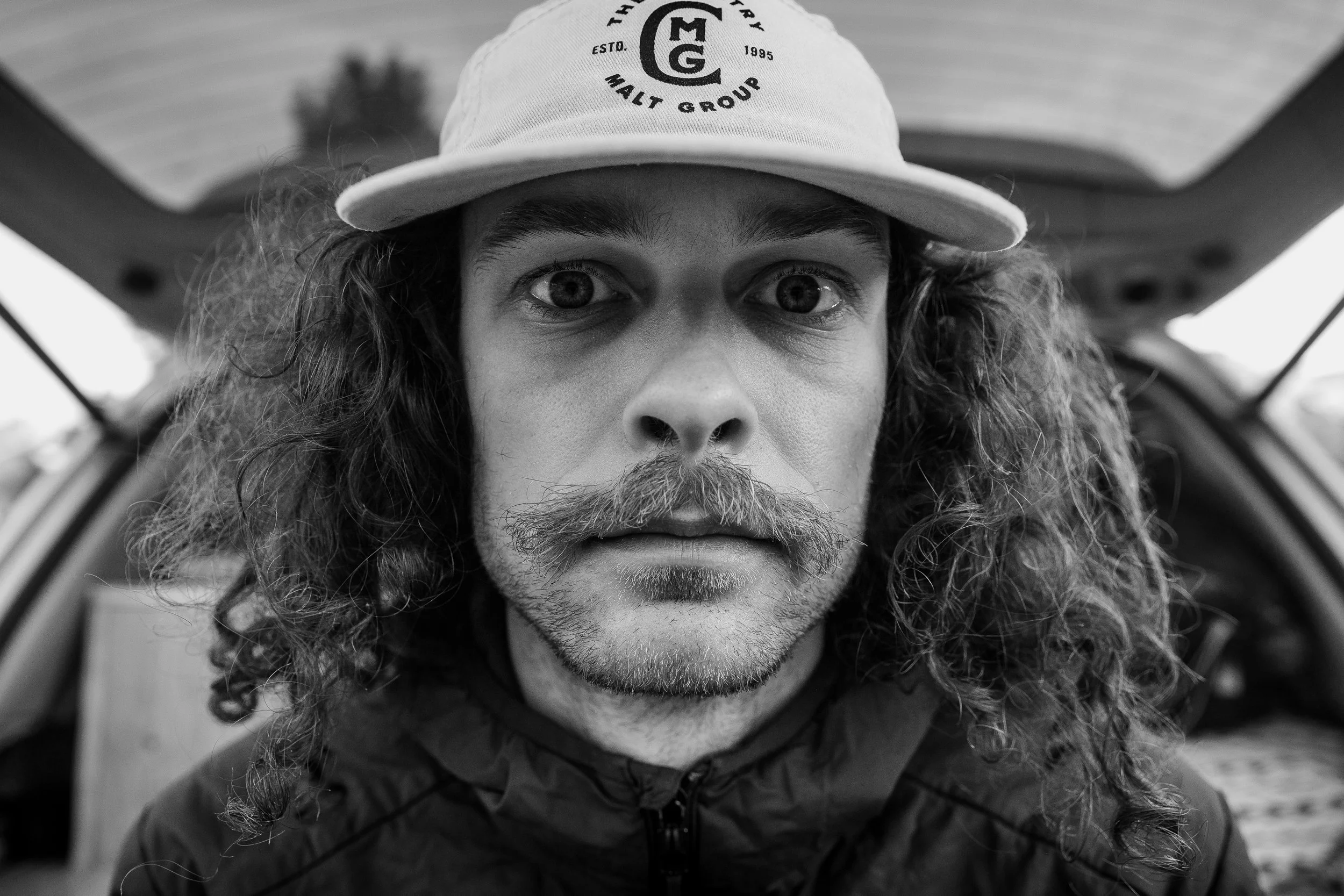 A black-and-white close-up portrait of a man with long, curly hair and a beard, wearing a baseball cap with the logo 'MG Malt Group' on it. He is looking directly at the camera with an intense expression, and the background appears to be outdoors.
