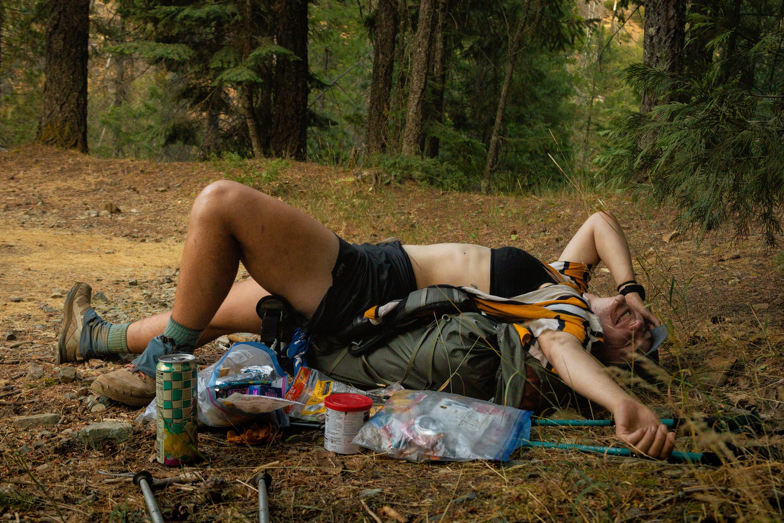 A person lying on their back on the ground in a forest, smiling, with camping or hiking gear around them.