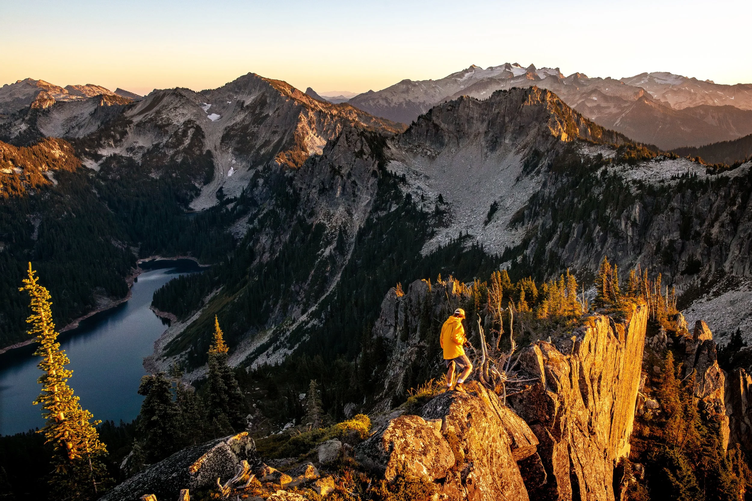 A mountain landscape at sunrise or sunset with rugged peaks, a lake, and a person in a yellow jacket walking on rocky terrain.