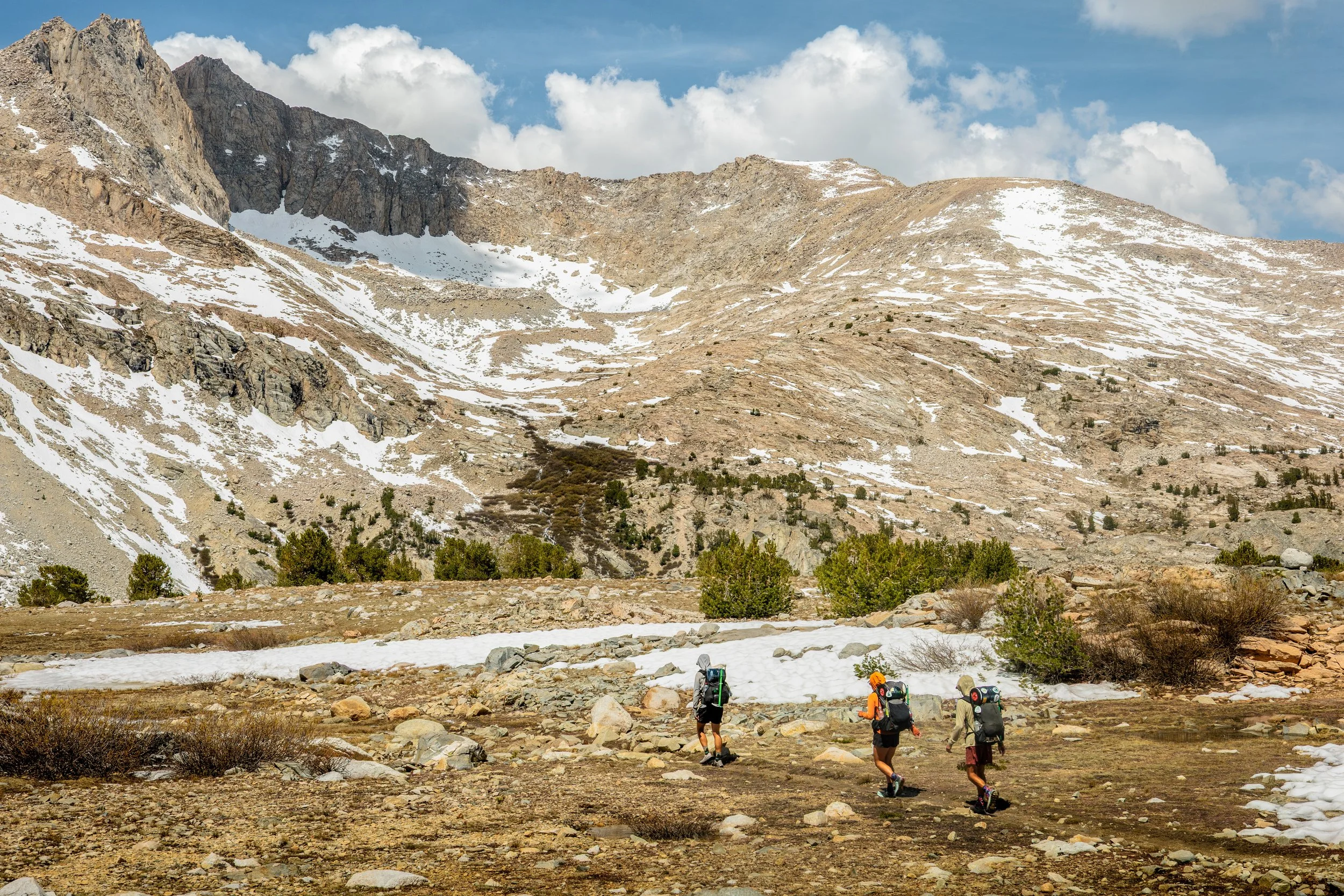 A group of hikers walking through a mountainous terrain with patches of snow, rocky slopes, and sparse trees, under a partly cloudy sky.