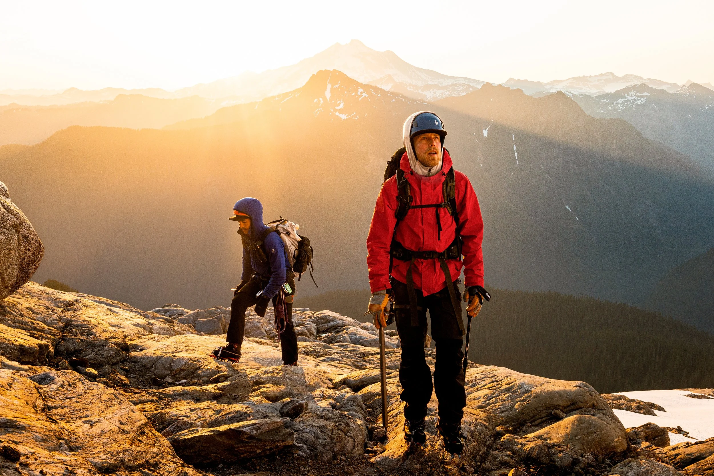 Two hikers, one in red and the other in blue, walking on rocky terrain at sunrise or sunset with mountain peaks in the background.