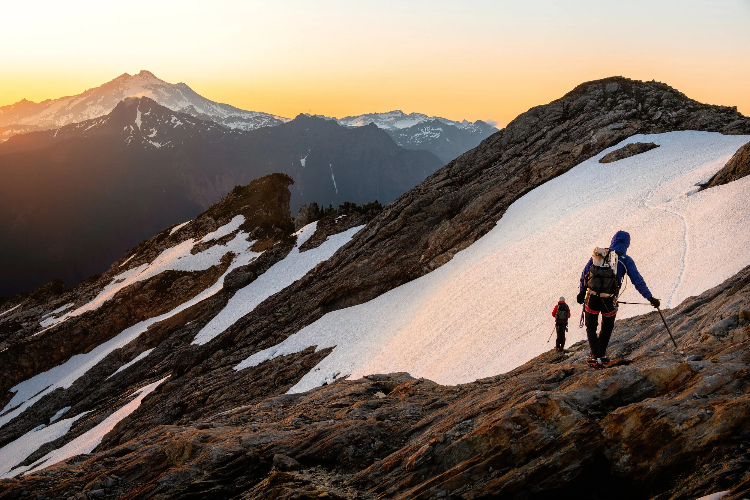 Two hikers climbing on a snow-covered mountain slope with rocky terrain, with a backdrop of snow-capped mountains and a sunset sky.