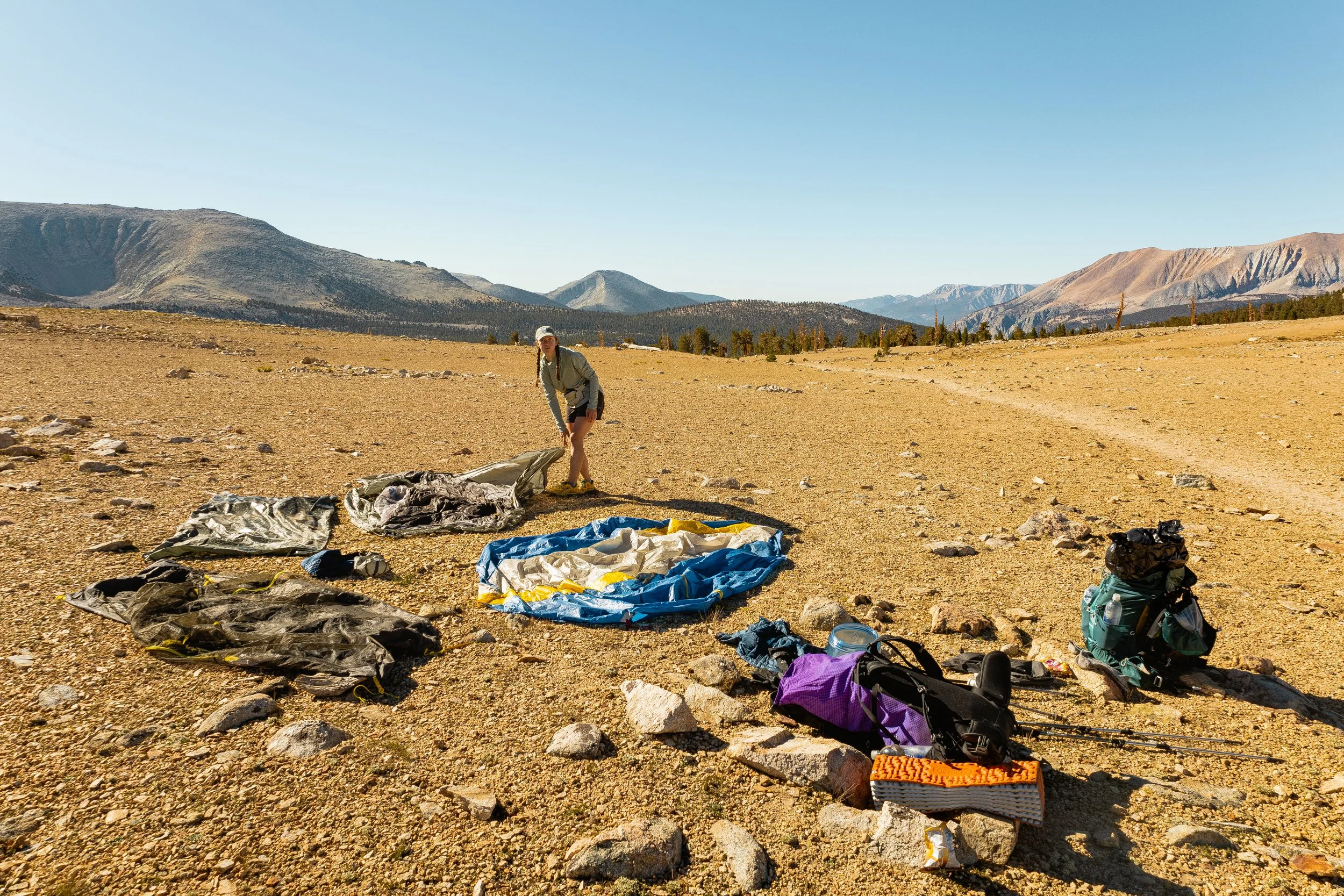 Person setting up camping gear on a rocky, open mountain landscape with distant mountains and a clear blue sky.