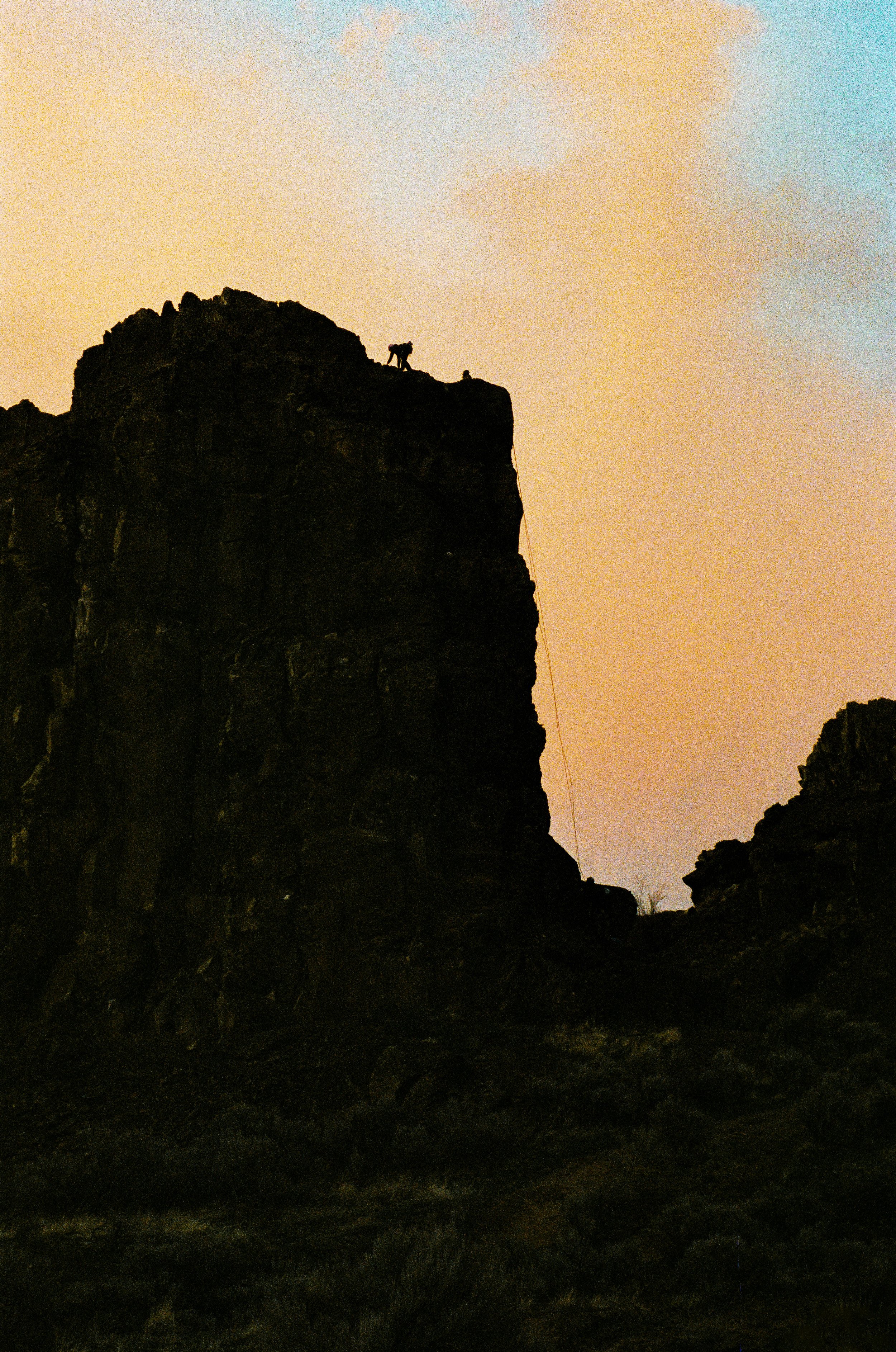 Silhouetted mountain landscape at sunset with a person rock climbing and a dog on top of the cliff.
