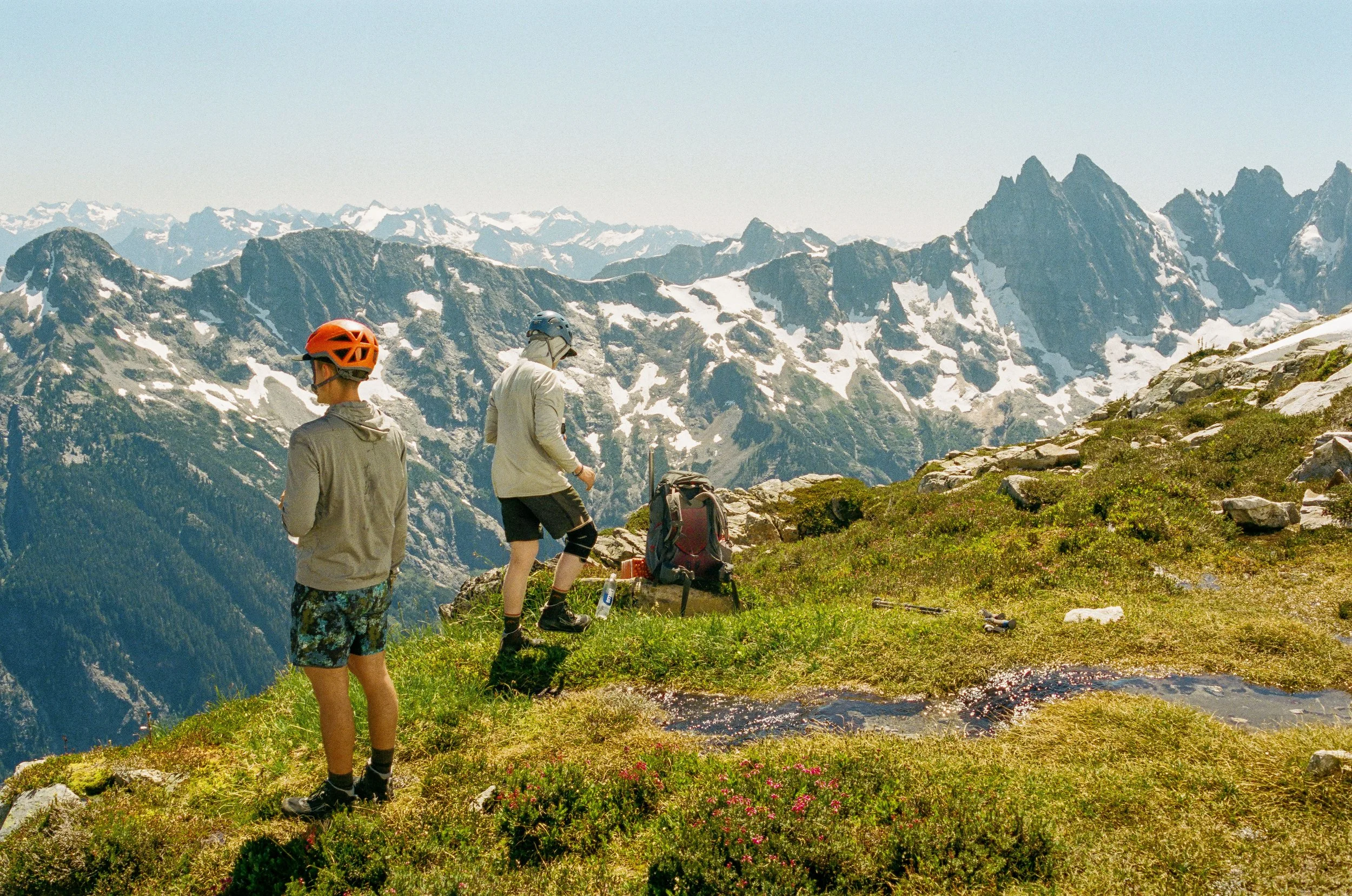 Two hikers with helmets and outdoor gear standing on a grassy, rocky mountain trail with snow-capped peaks in the background.