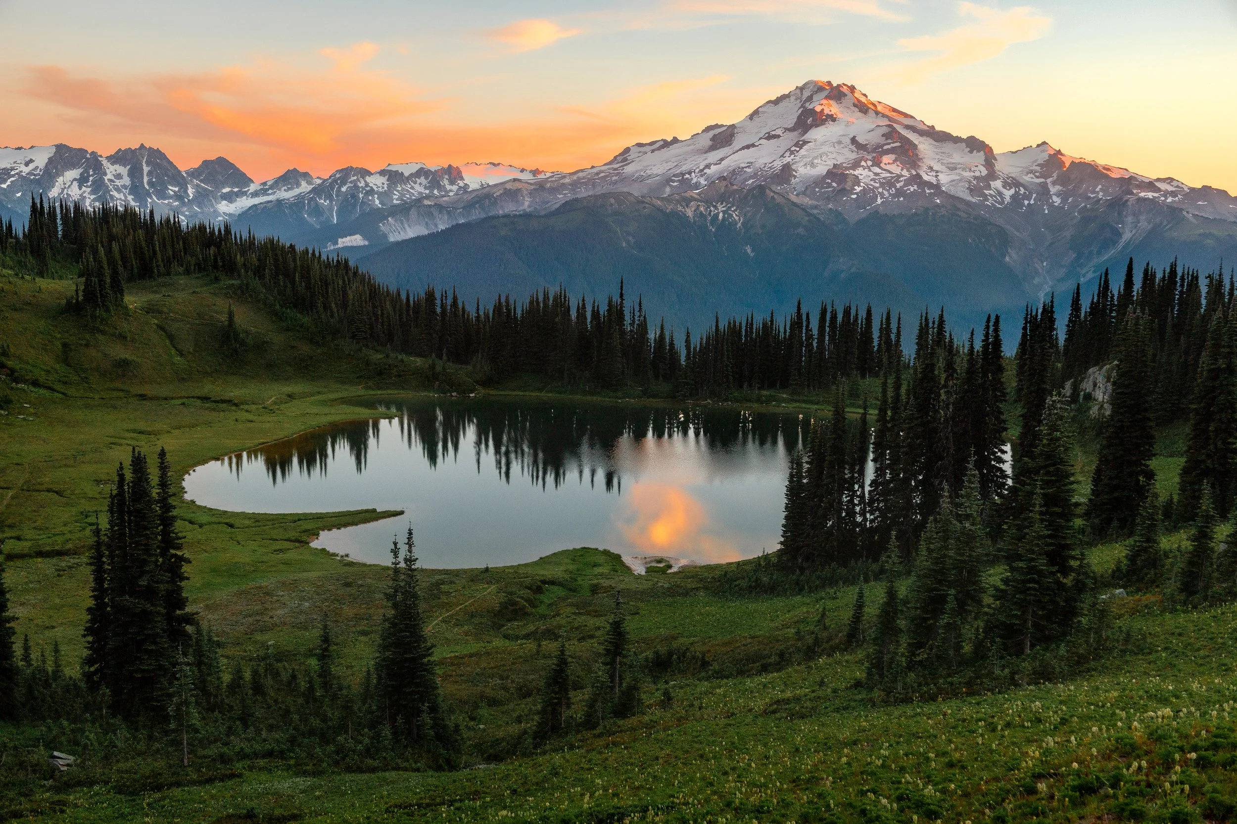 Snow-capped mountain peaks at sunset with a lake reflecting the sky and forested landscape in the foreground.