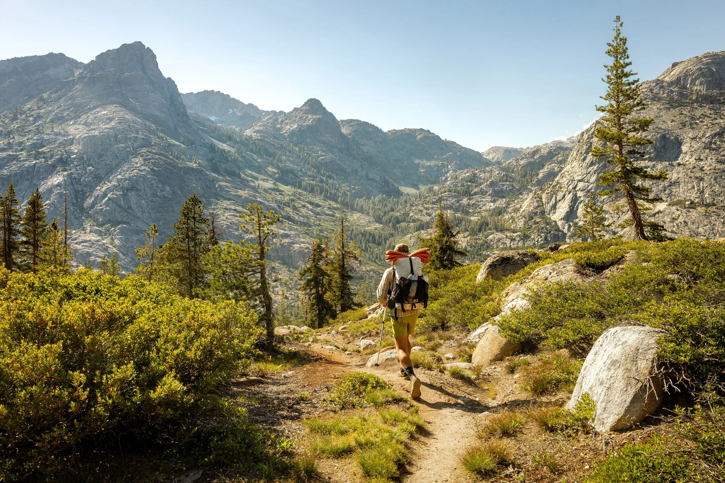 A person wearing a hat, hiking gear, and shorts walks along a trail in a mountainous forested area. The background features large rocky mountains under a clear sky.