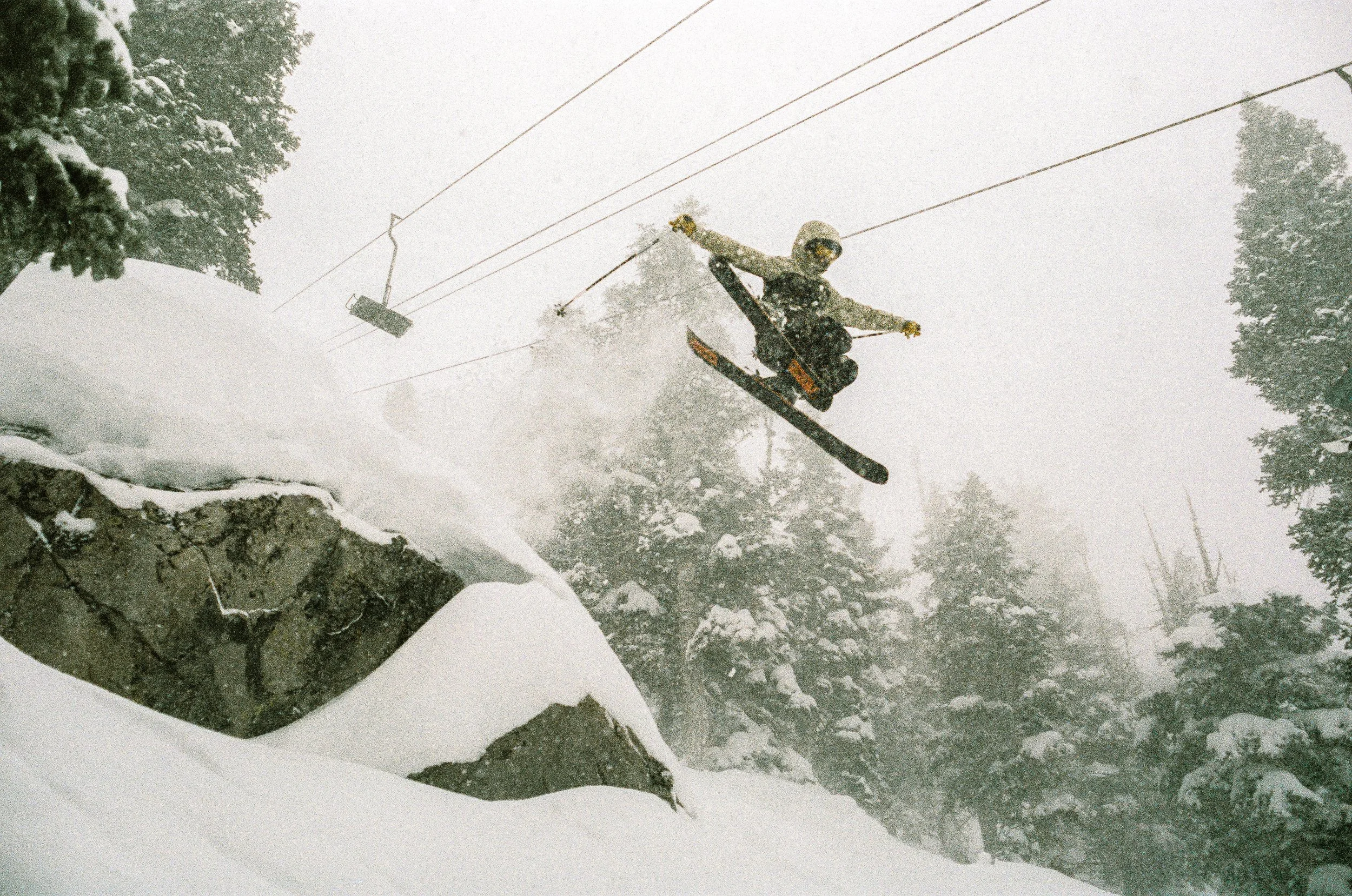 A person in ski gear, including a helmet and goggles, jumps off a snow-covered ledge in a snowy, forested mountainous area during a snowstorm.
