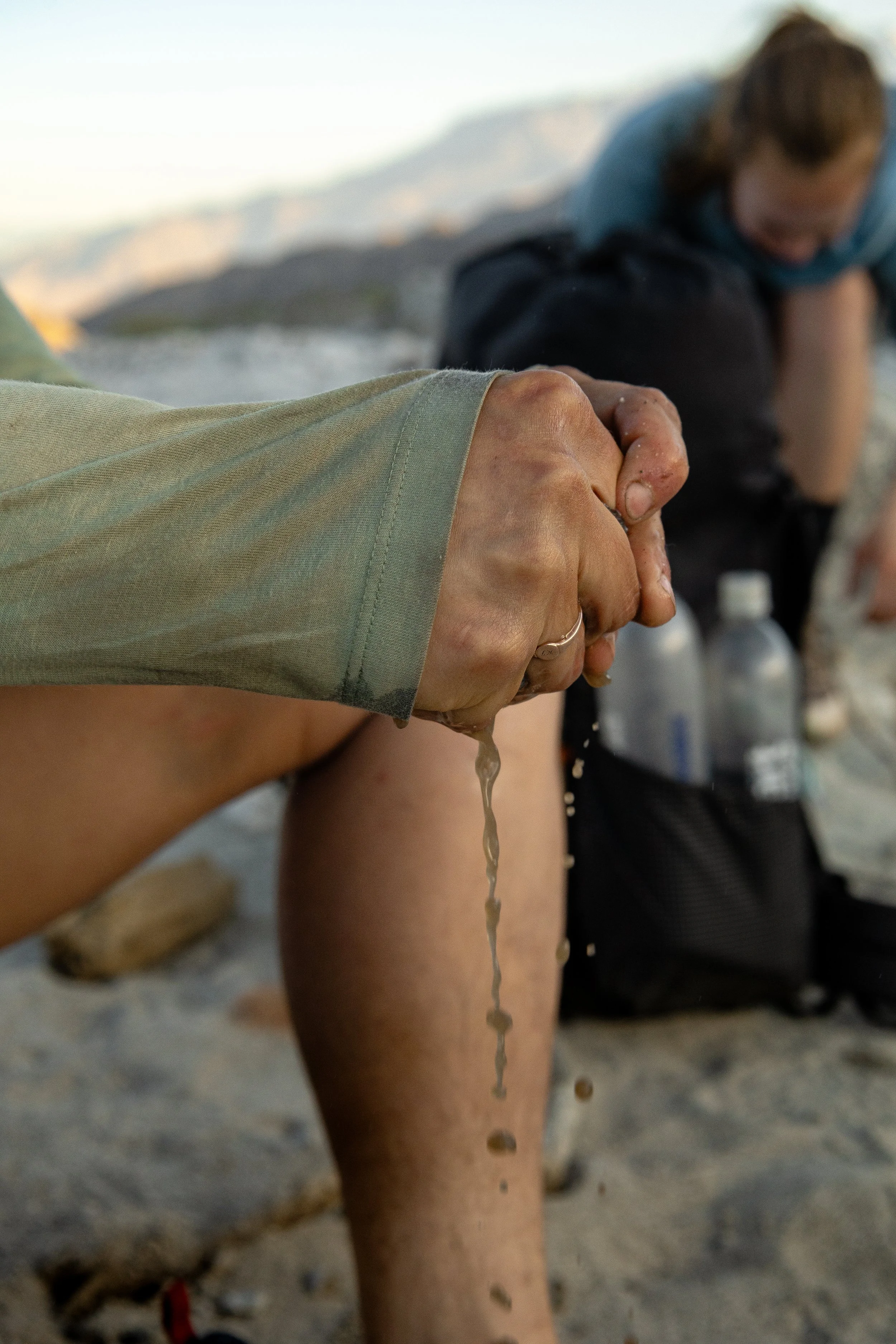 A person with sandy hands holding water from a bottle, with another person in the background sitting on a sandy surface, surrounded by water bottles and outdoor gear.