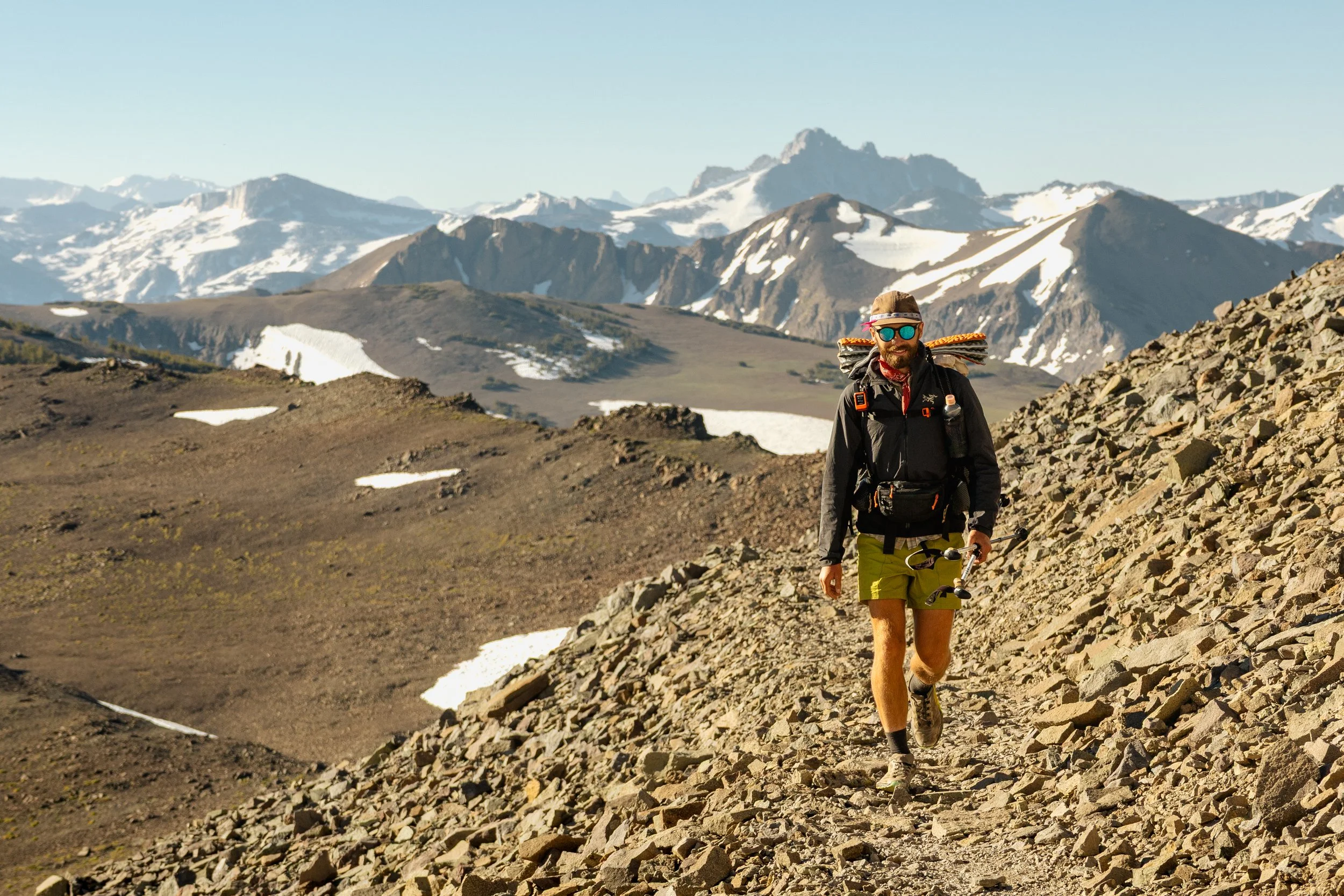 A man hiking in a mountainous area with snow patches, wearing sunglasses, a hat, and outdoor gear, walking along a rocky trail with mountain peaks in the background.