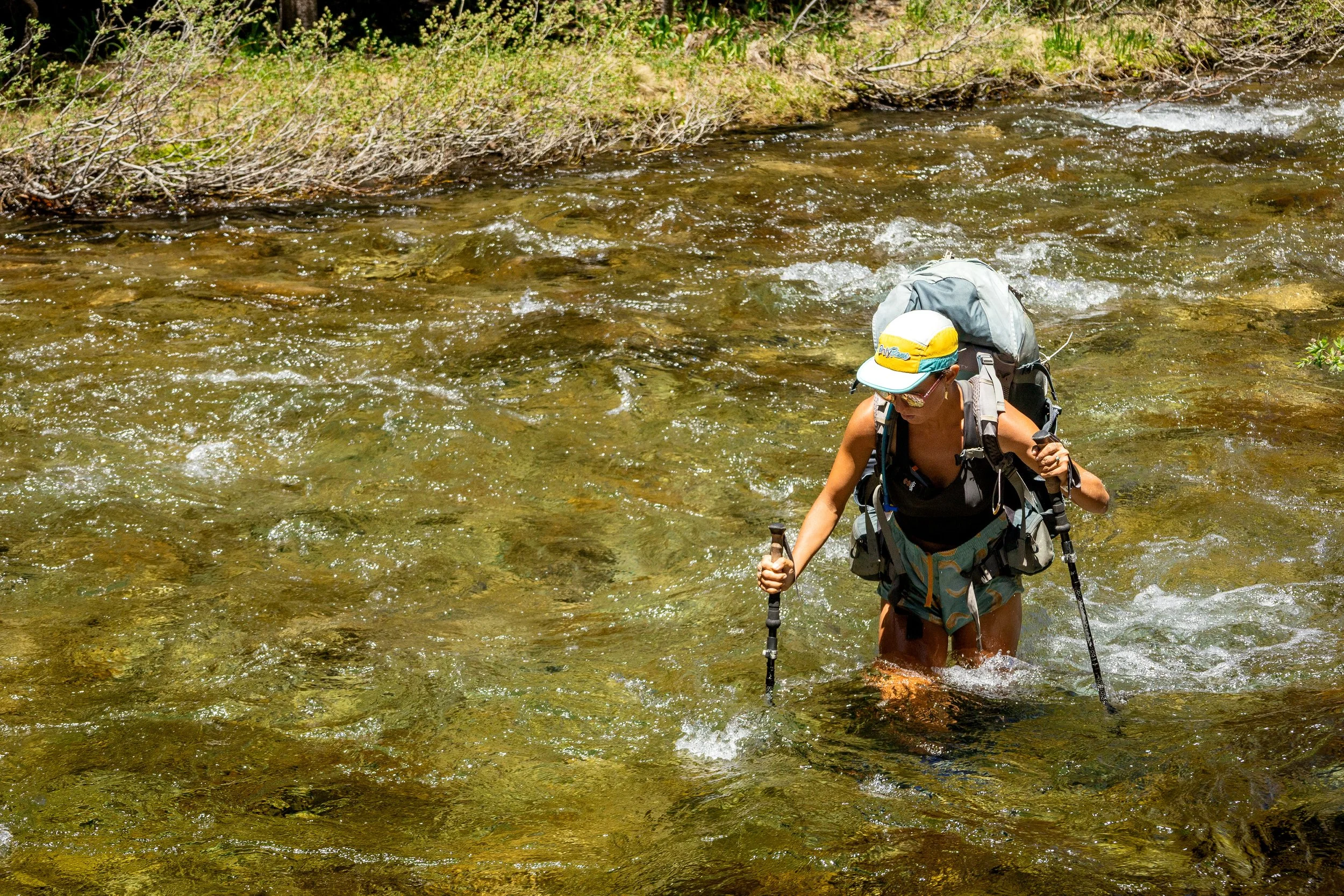 Hiker crossing a shallow stream in a forested area, wearing a hat, sunglasses, and a backpack, using trekking poles.