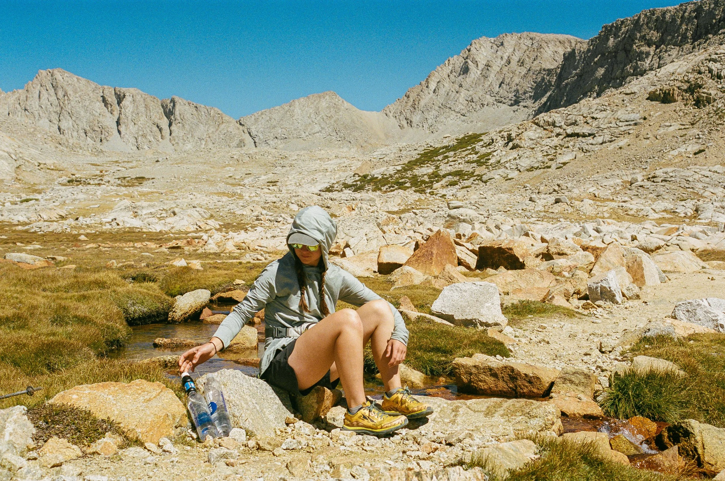 A person sitting on rocks by a small stream in a mountainous landscape with rocky peaks and a clear blue sky.
