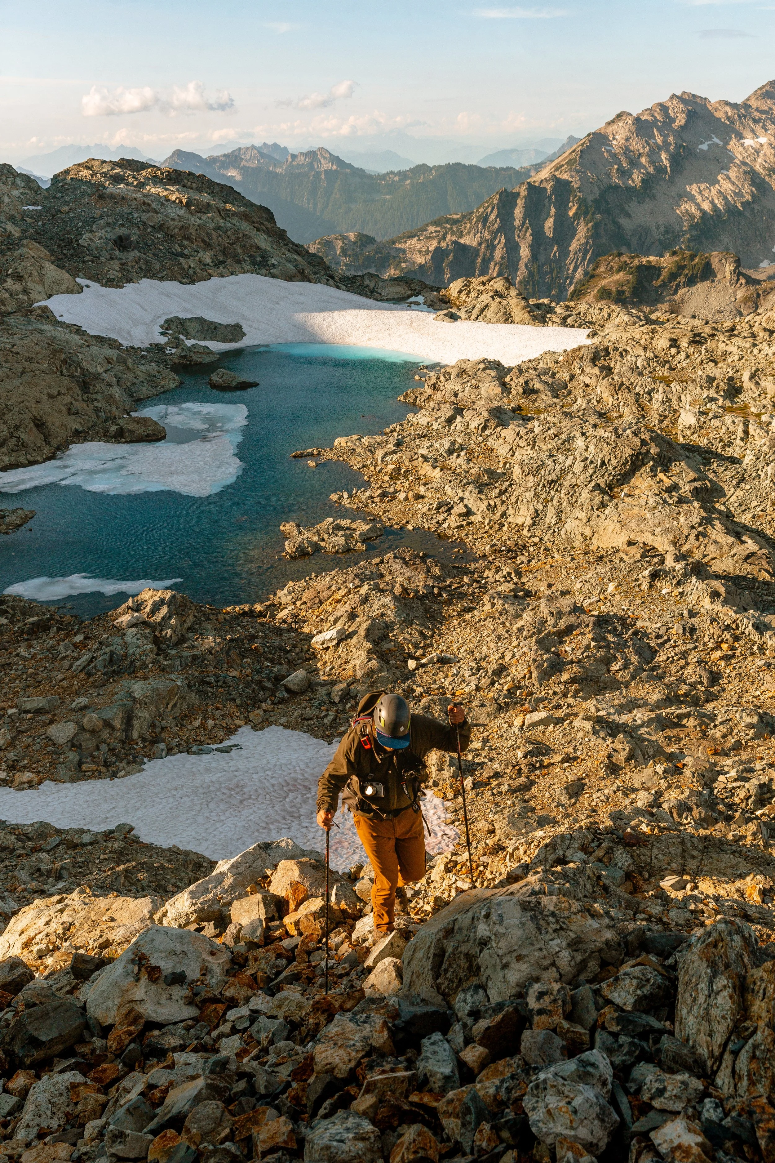 A person wearing a helmet, sunglasses, and outdoor gear climbing a rocky mountain trail with snow patches in the foreground and a mountain lake in the background.