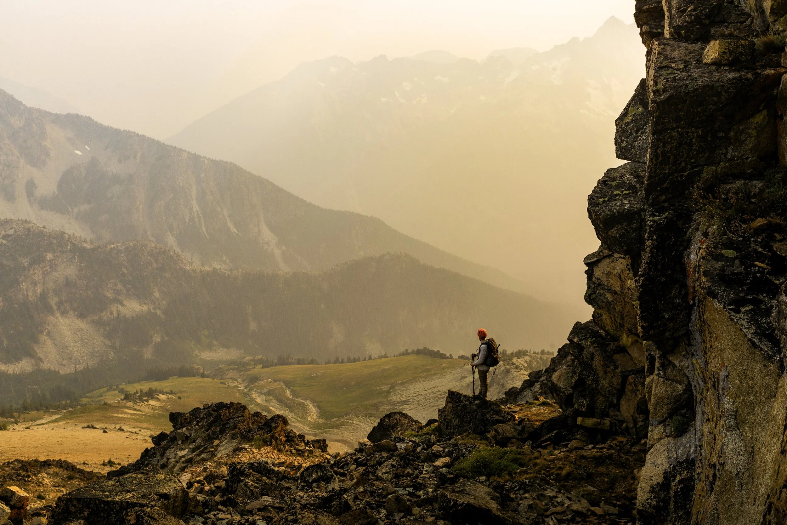 Hiker with red helmet and backpack standing on rocky terrain beside a large stone cliff with mountain range in the background during sunset.