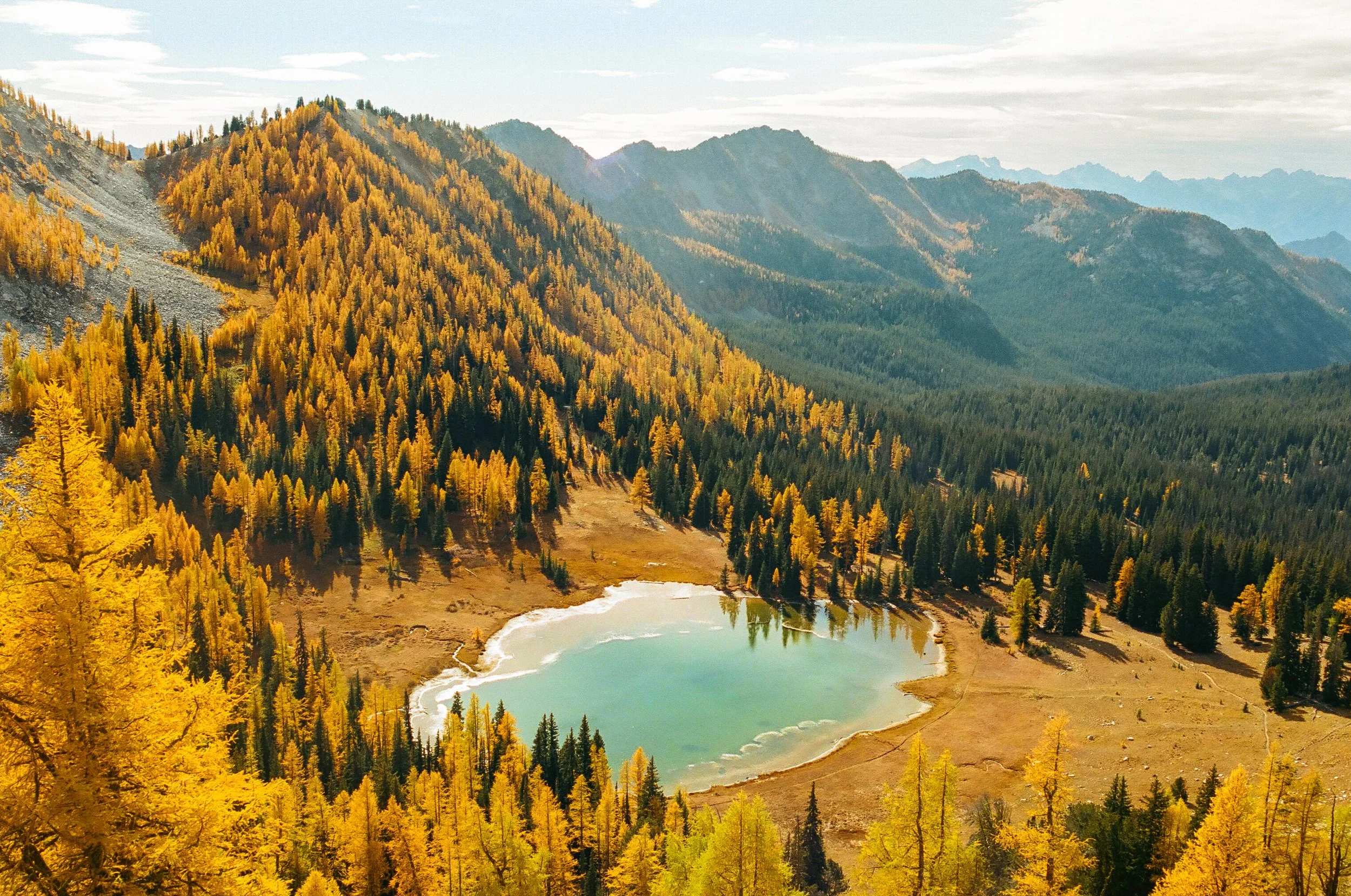 A scenic landscape of a mountain range with dense yellow and green trees, a small lake in the foreground, and a partly cloudy sky.