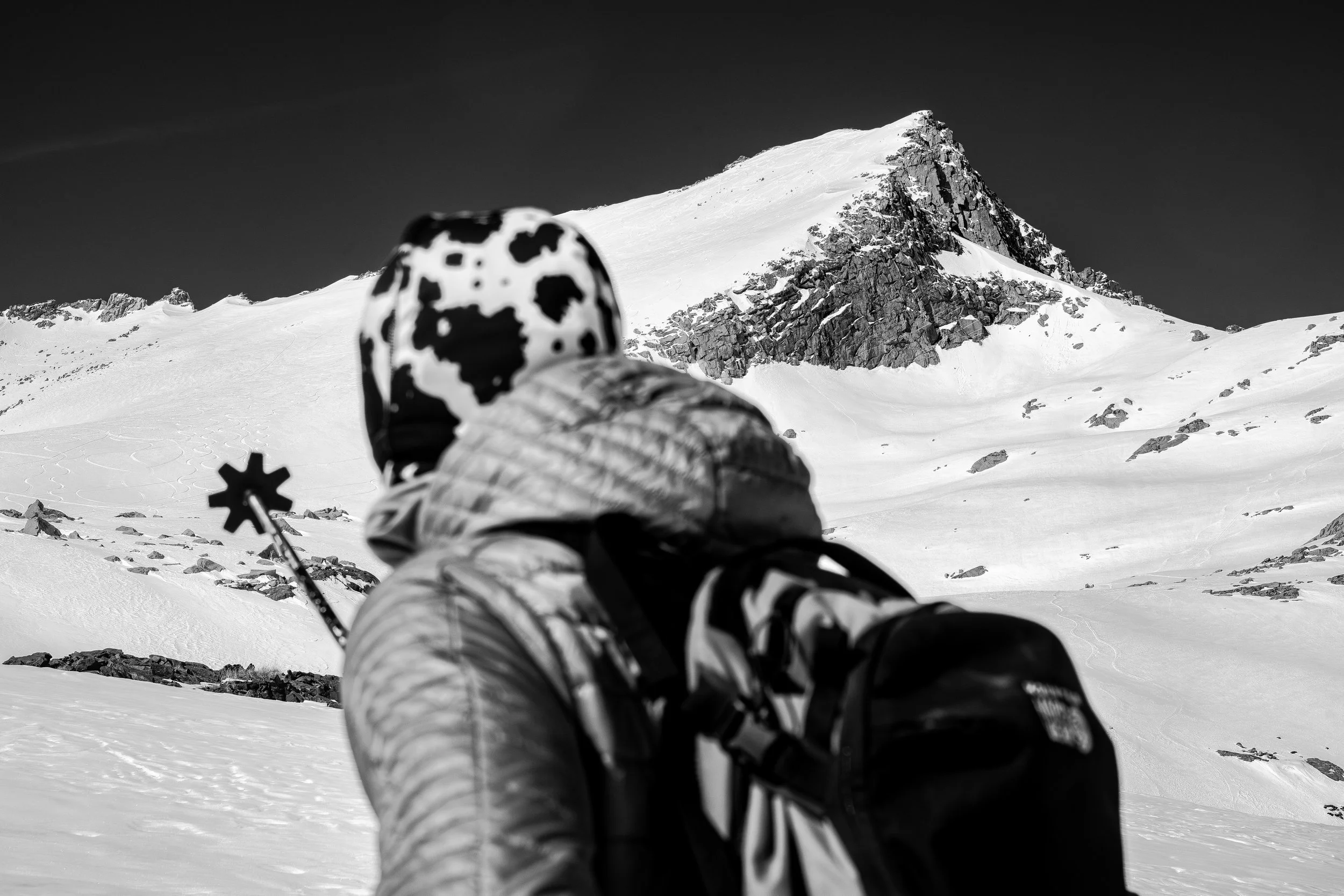A person in winter clothing lying on their back on snow, holding ski poles, with snow-covered mountains in the background.