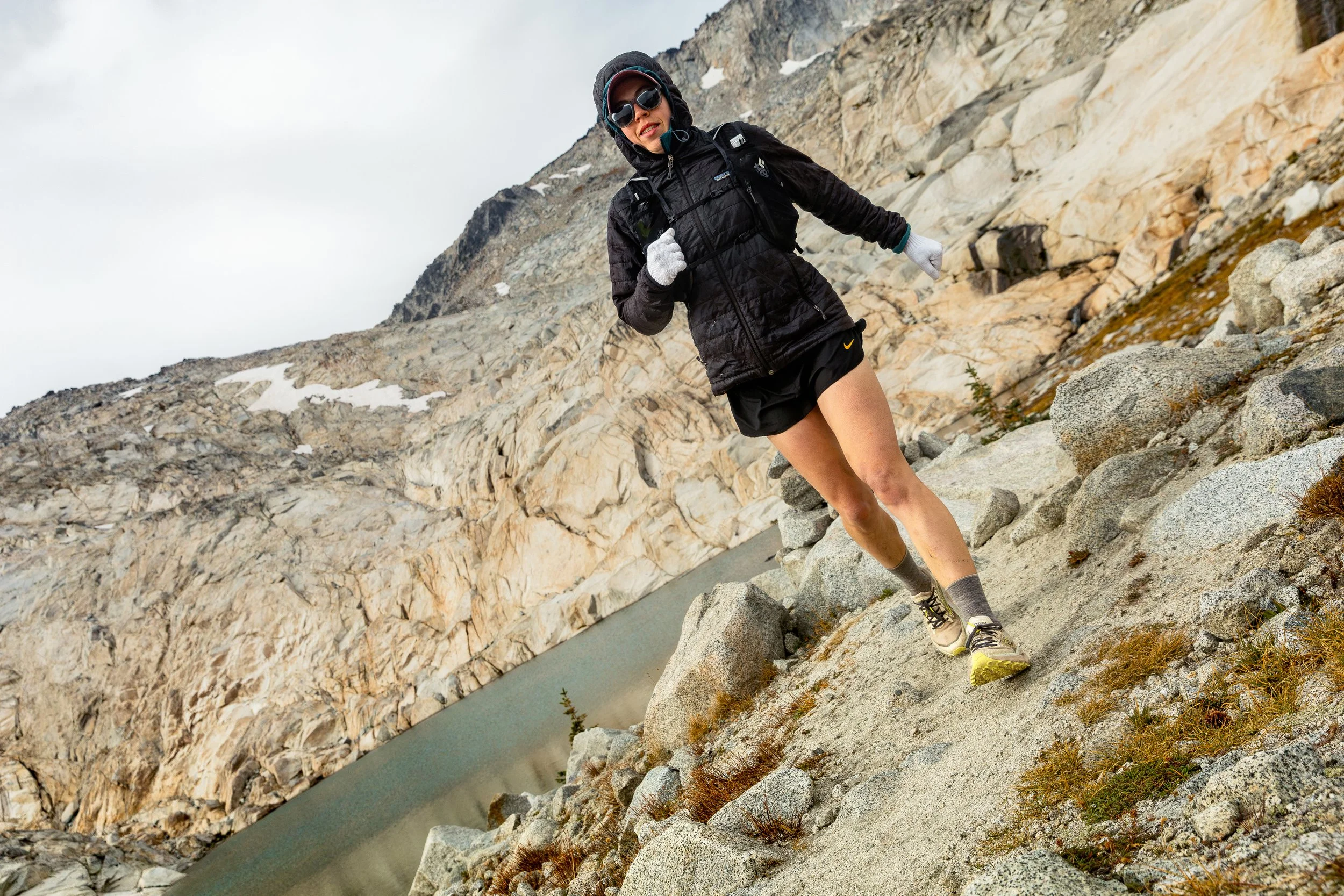 A woman trail running on rocky terrain near a mountain lake, dressed in athletic gear, wearing sunglasses, a black jacket, shorts, and running shoes.