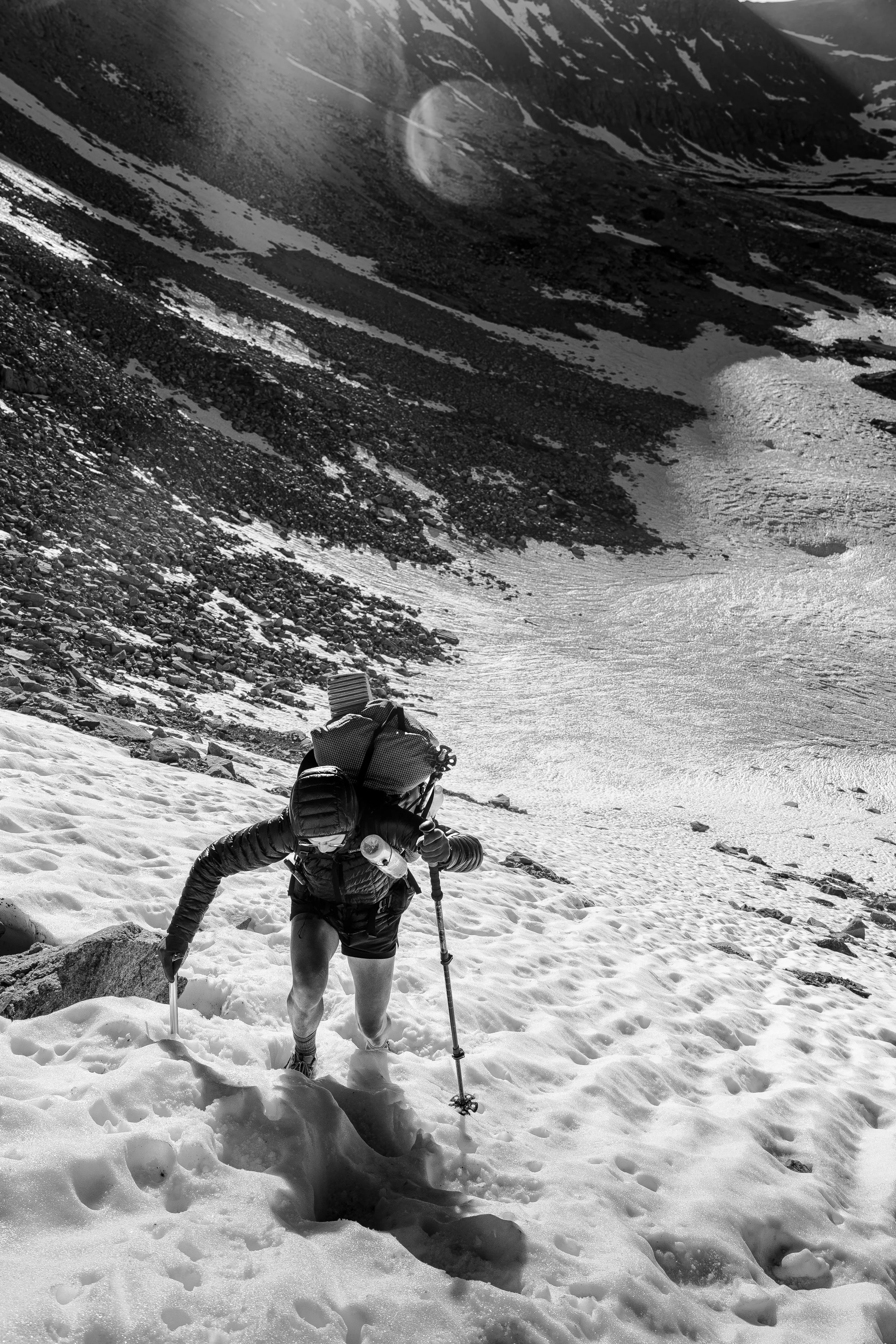 A person wearing outdoor gear climbing snow-covered terrain with ice axes in a mountainous landscape with rocky slopes and snow patches.