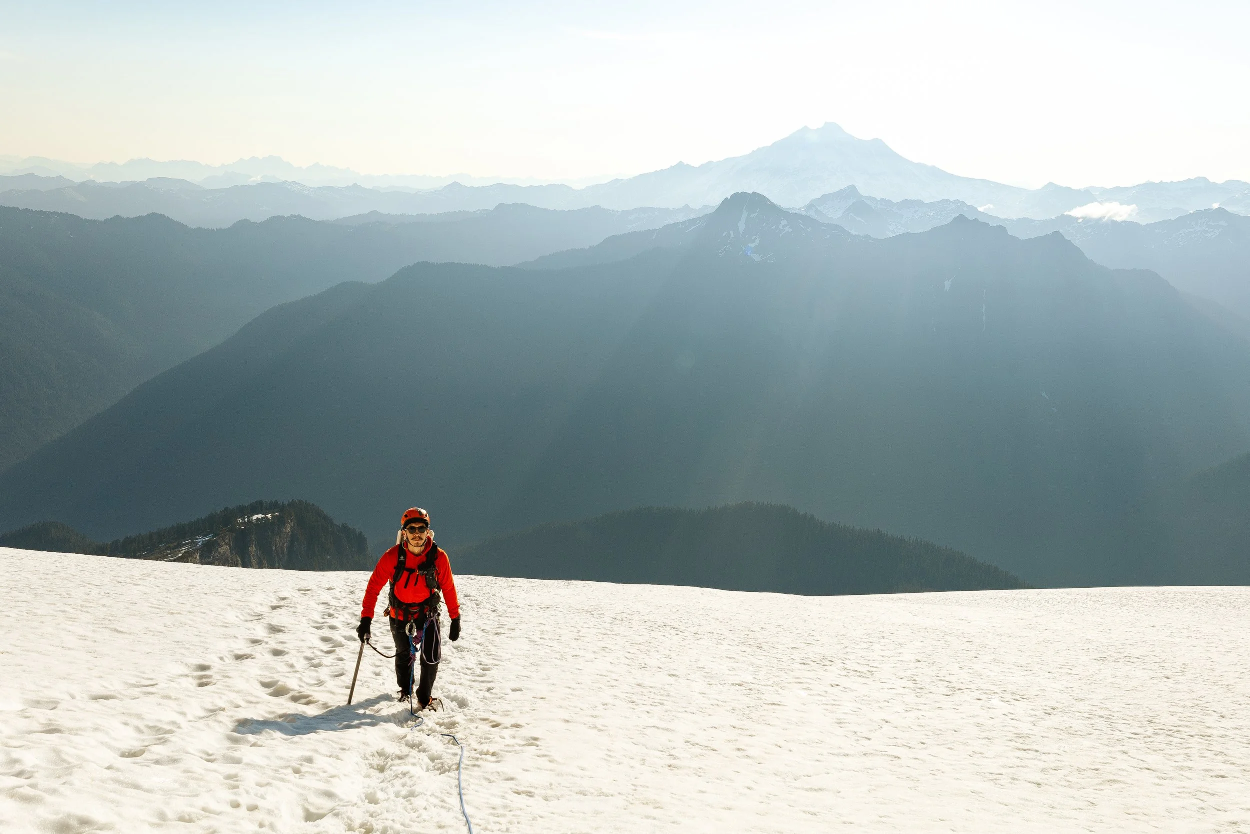 A male hiker wearing a red jacket, black pants, and a red helmet walking in the snow on a mountain, with mountain ranges and a partially snow-capped peak in the background.