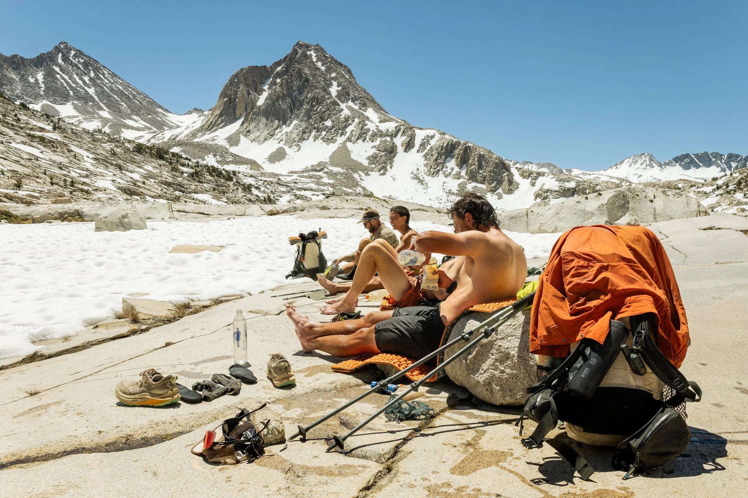 Three people relaxing on rocks in a snowy mountain landscape with rugged peaks and clear blue sky, with hiking gear and water bottles nearby.
