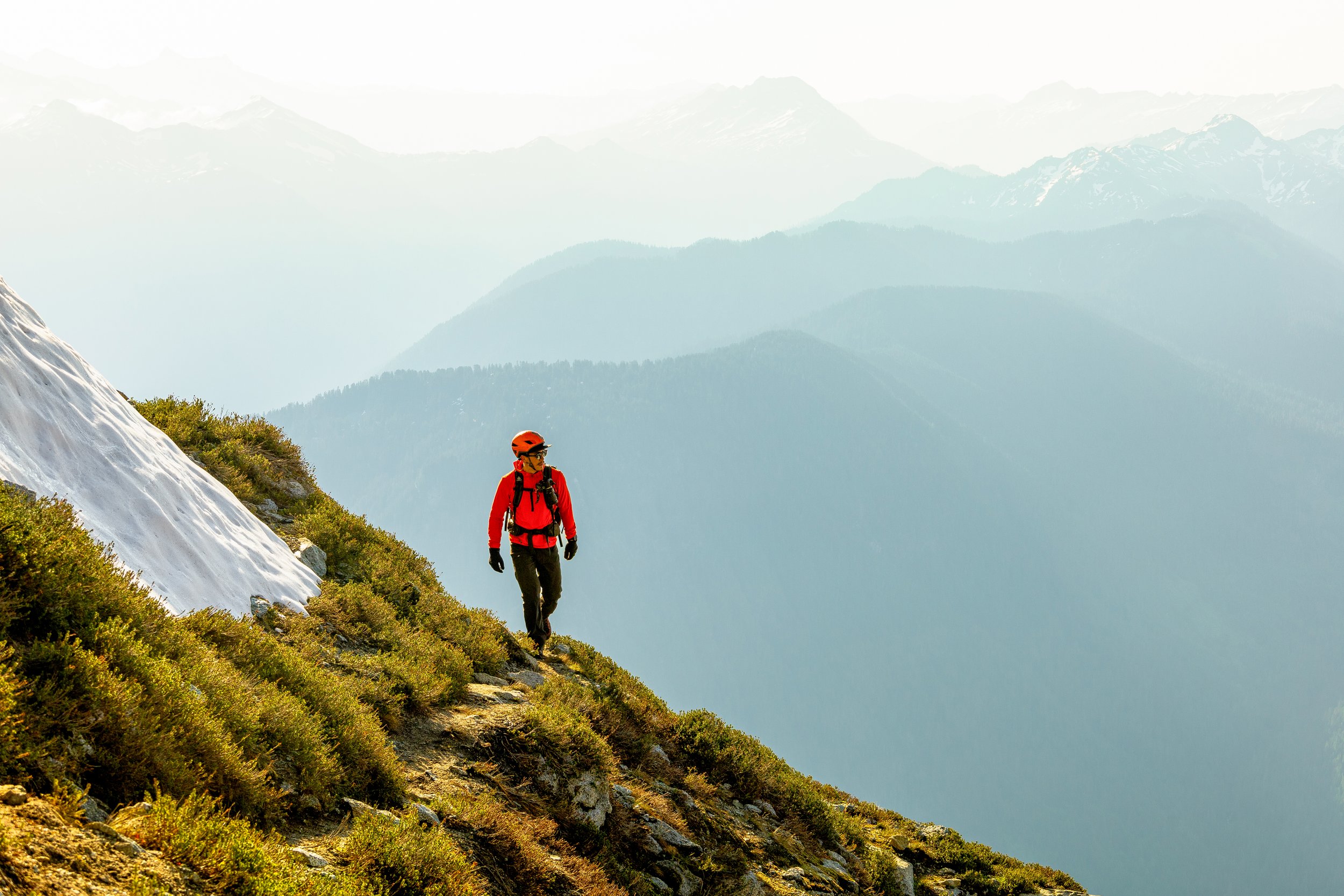 A hiker wearing a red jacket and helmet walking on a mountain trail with snow and green shrubs, against a backdrop of multiple mountain ranges under a clear sky.
