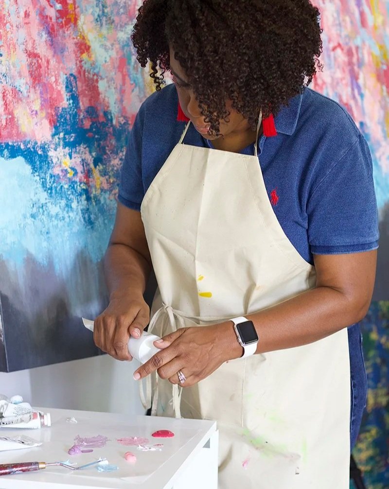 Woman with curly hair wearing a blue polo shirt and a white apron, using a piping tool, in front of a colorful, abstract painted background.