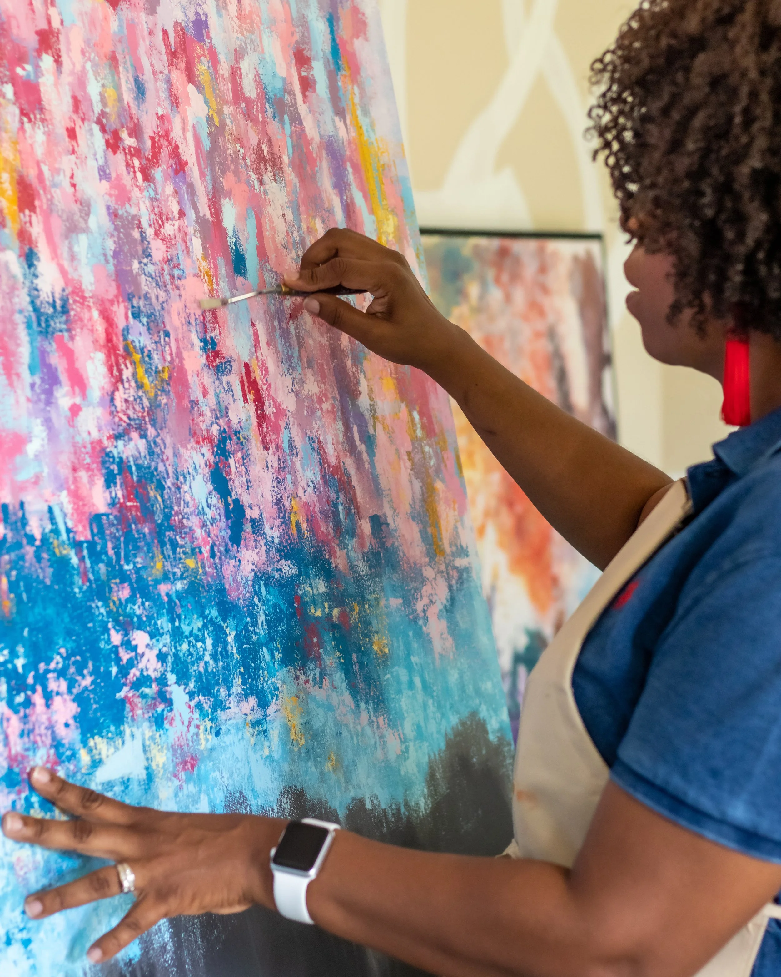 A person with a dark skin tone painting an abstract artwork with bright colors using a palette knife on a large canvas.
