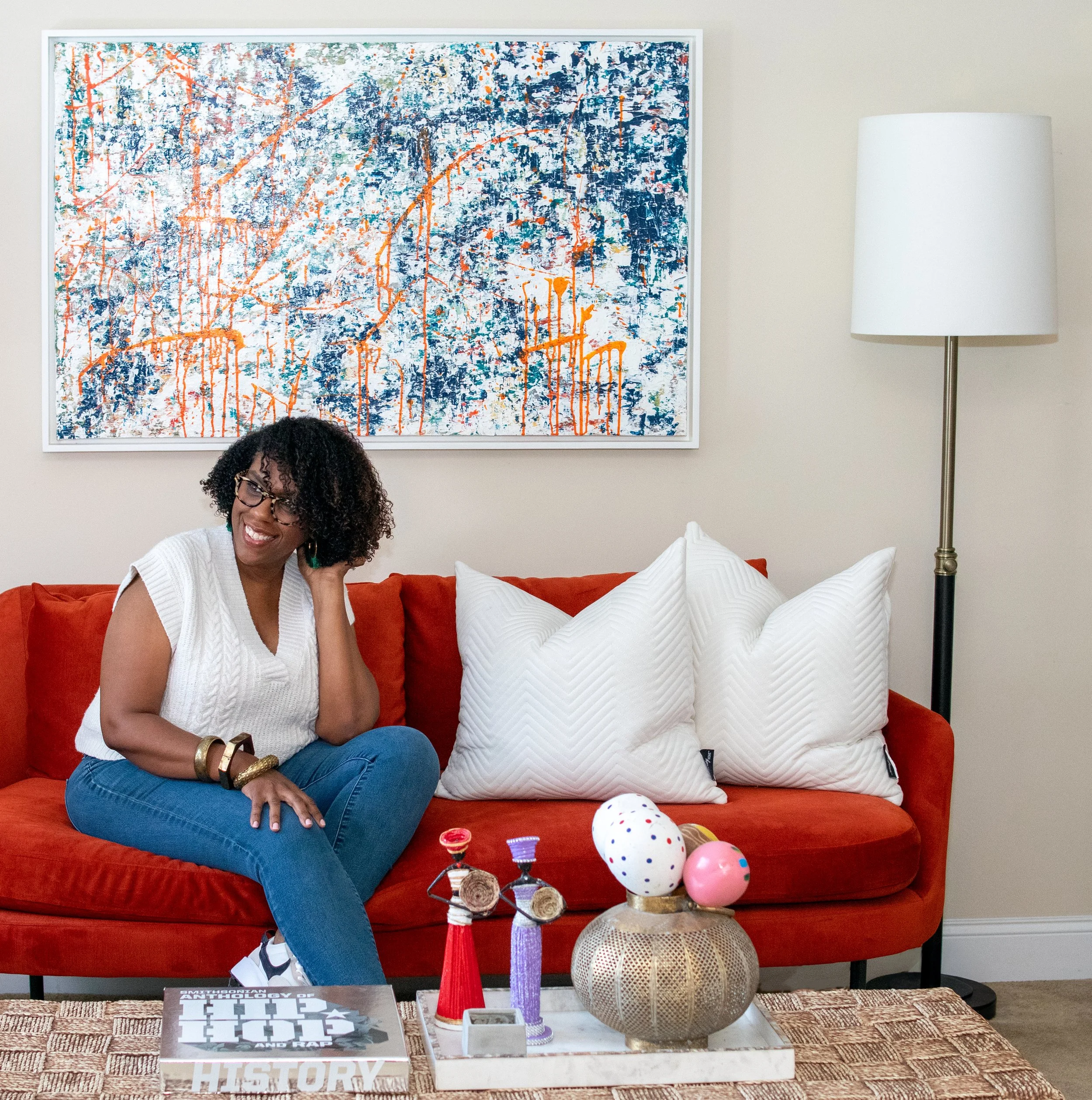 The artist, Kathleen Mapson, with curly hair and glasses smiling at a table with decorative objects, in a room with a large colorful abstract painting on the wall behind her, and a white lamp to the right.
