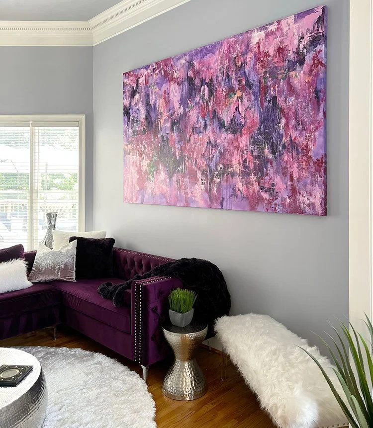 Living room with a purple velvet sectional sofa, white furry pillows, abstract pink and purple painting on the wall, white shaggy rug, silver side table with a potted plant, and a window with blinds.