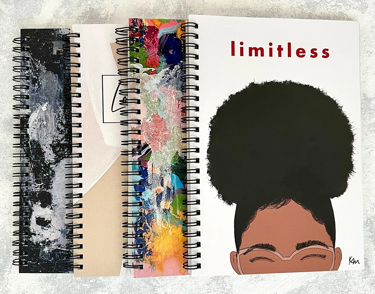 Three spiral-bound notebooks with abstract and colorful covers, the rightmost notebook features an illustration of a smiling girl's face with natural hair and the word "limitless" in red.