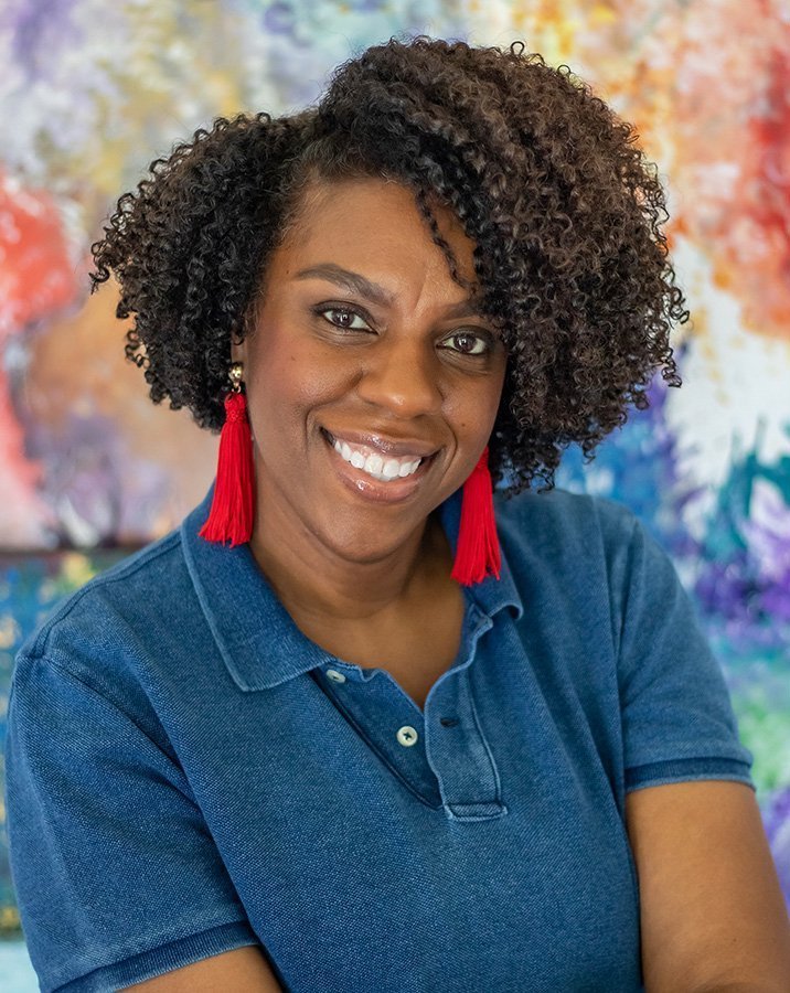 A woman with curly hair wearing a blue polo shirt and red tassel earrings, smiling against a colorful blurred background.