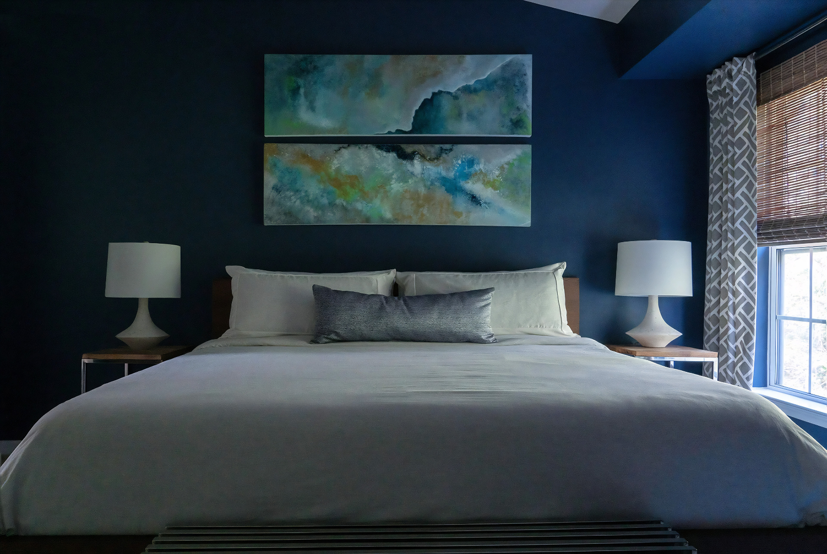 A bedroom with a dark blue accent wall, featuring two modern table lamps on nightstands, a large bed with white bedding and gray pillows, abstract landscape paintings above the bed, and a window with patterned curtains letting in natural light.